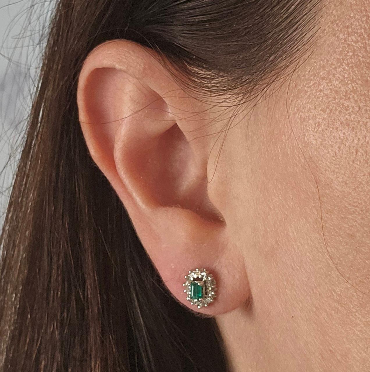 18 Karat White Gold Vintage Stud Earrings Featuring 2 Emerald Cut Emeralds Totaling Approximately 0.20 Carats Surrounded by 28 Round Single Cut Diamonds of SI Clarity and G/H Color Totaling 0.25 Carats. Pierced Post with Friction Backs. Finished