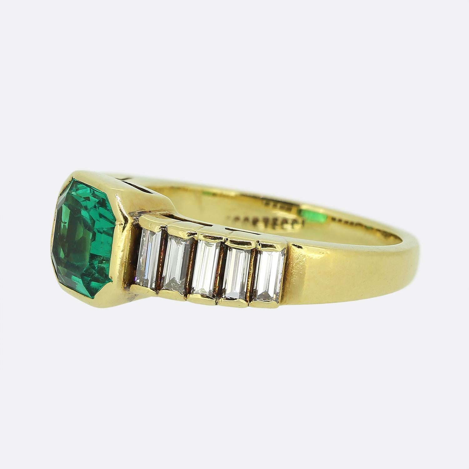 Here we have a exquisite 18ct yellow gold ring featuring a striking green emerald at its centre. This vibrant principal stone is set in a bold bezel setting whilst being flanked on either side by a single row of baguette-cut diamonds, channel-set