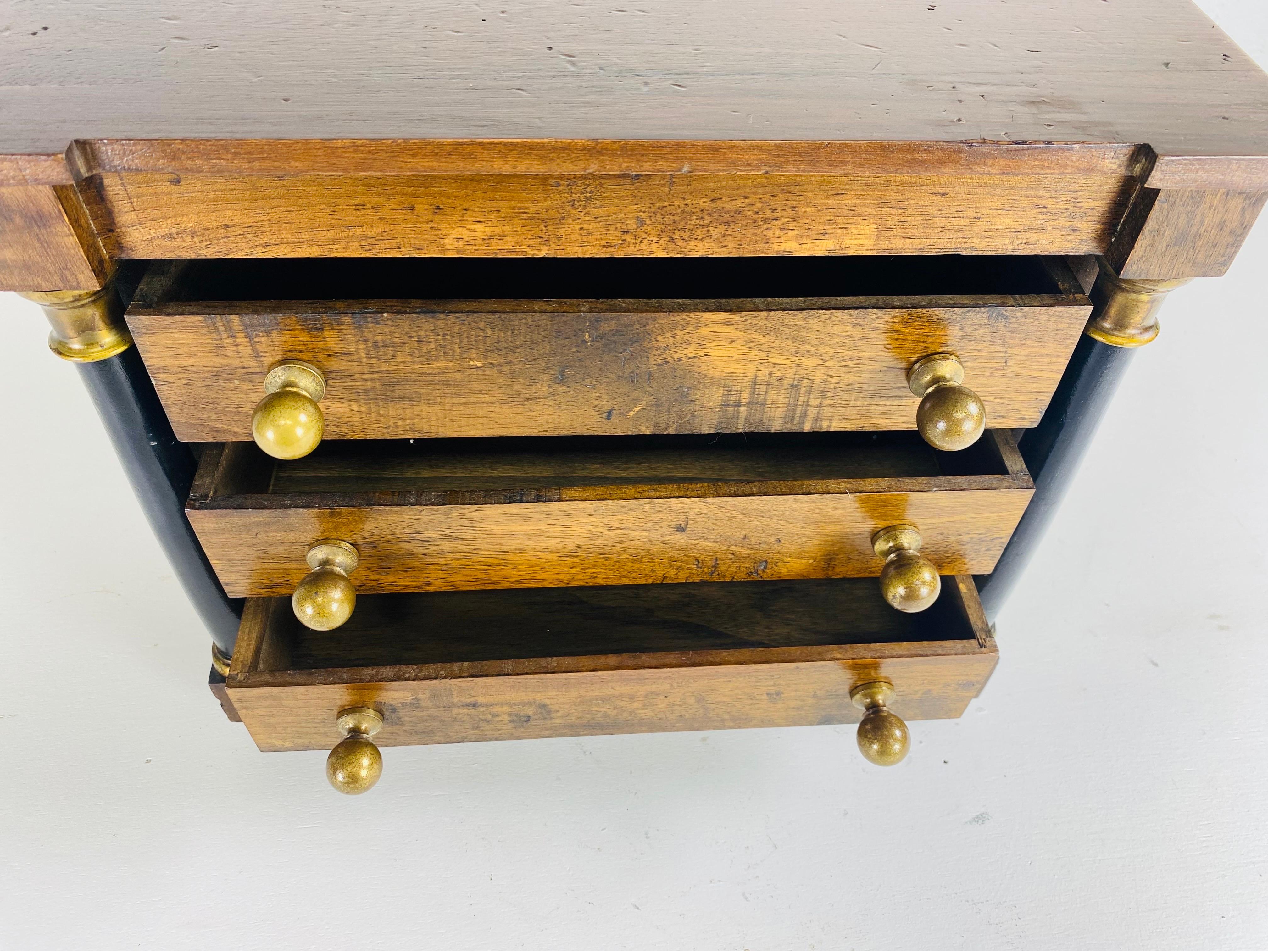 This is a Vintage empire style jewelry or trinkets chest of drawers. This chest of drawers features its original solid brass hardware. This diminutive chest of drawers is made of dark walnut with two columns on either side in black. This jewelry