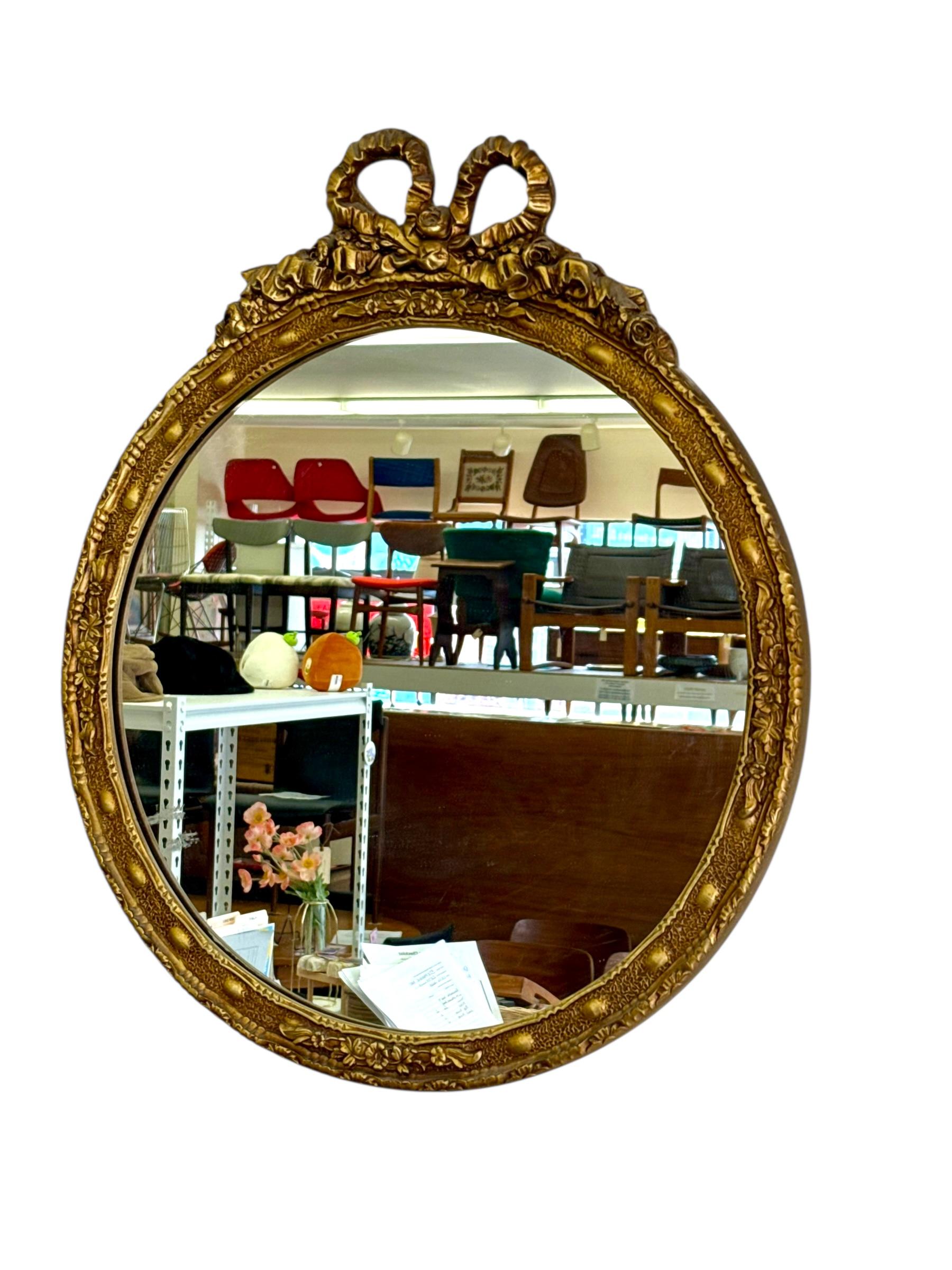 Ce miroir rond vintage est parfait pour une entrée ou au-dessus d'un lavabo dans une salle de bain. Le miroir est d'origine, avec quelques petites imperfections. Le cadre est doré et ne présente aucune perte de dorure. La partie supérieure est ornée