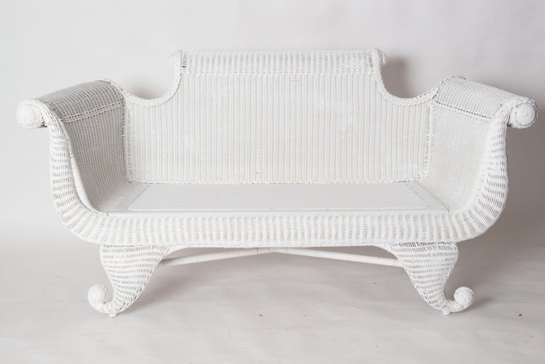 A whimsical wicker loveseat in the Empire style painted white. All white wicker/rattan with a new caned seat. Curled feet, rolled arms, and curled back. Quite a conversation piece.