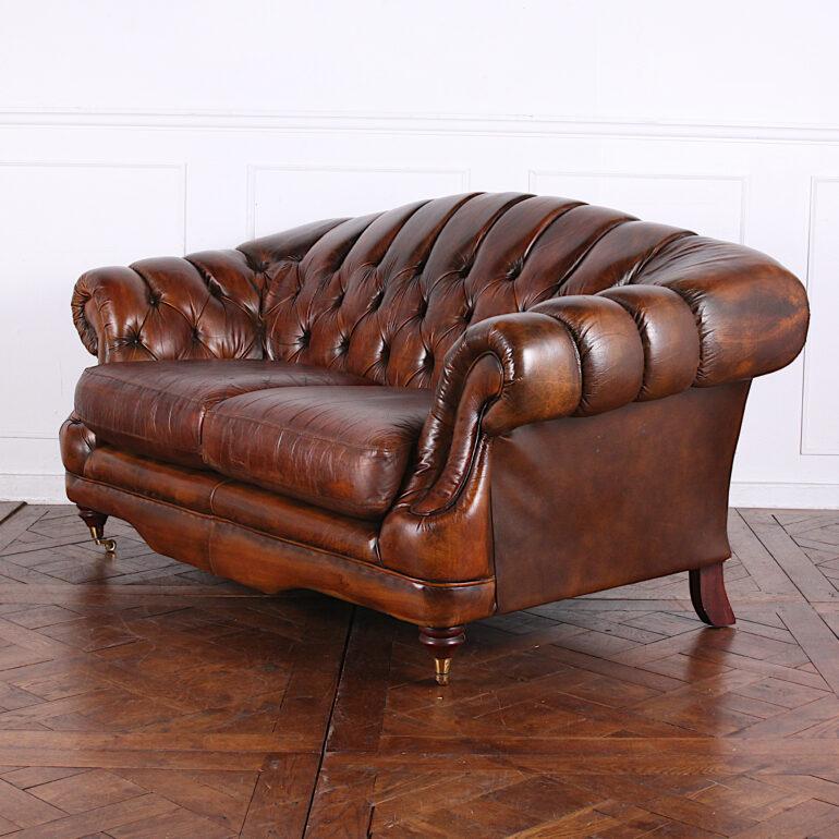A vintage English camel-back sofa in button-tufted leather with turned front feet with brass castors. Leather seat cushions with some wear but really just 'patina' and still serviceable. Classic warm brown leather colour.