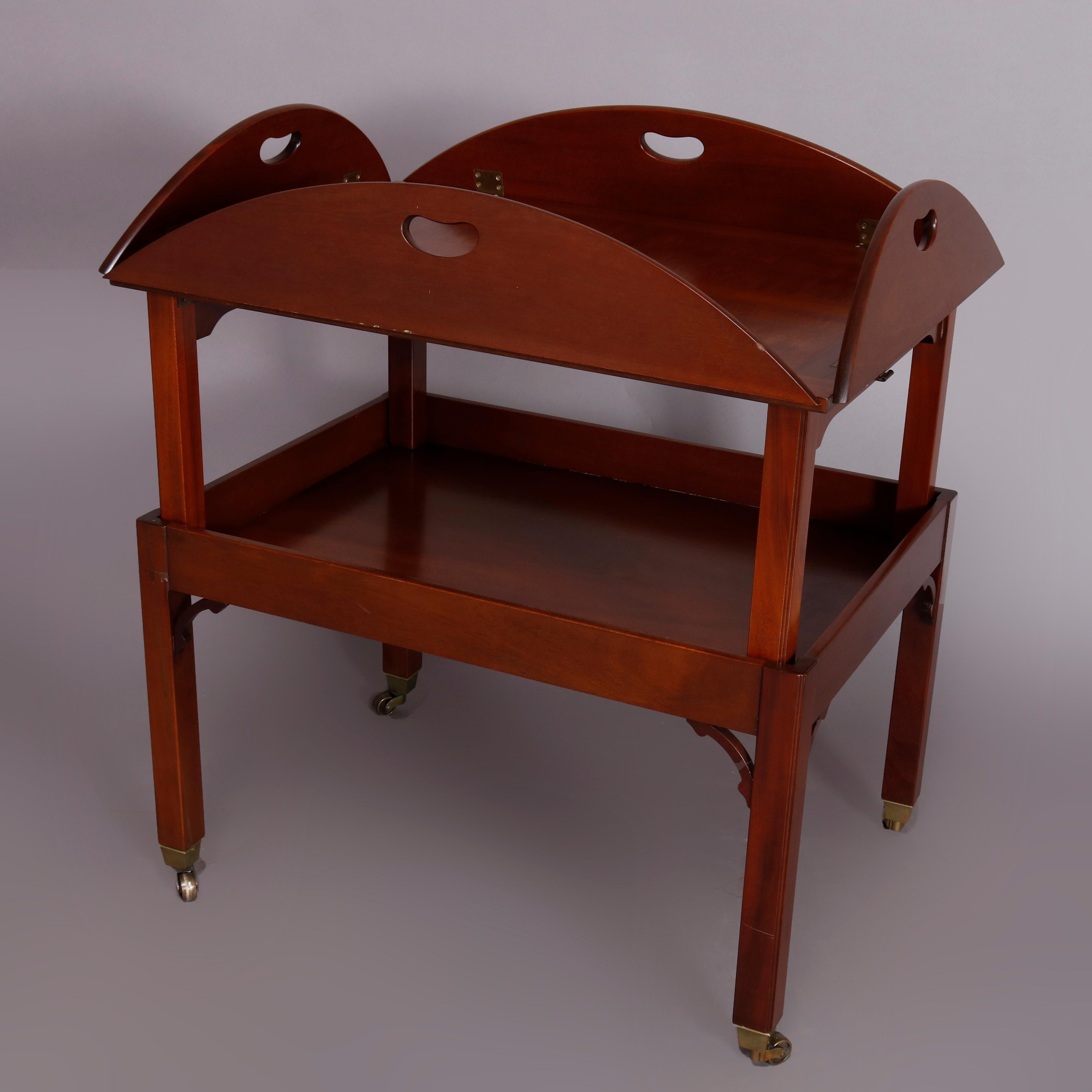 A vintage English George III tea table by Kittinger offers mahogany construction with inlaid top having handled and hinged flip-up sides surmounting lower shelf and raised on straight and square legs with shaped corbels and terminating in casters,