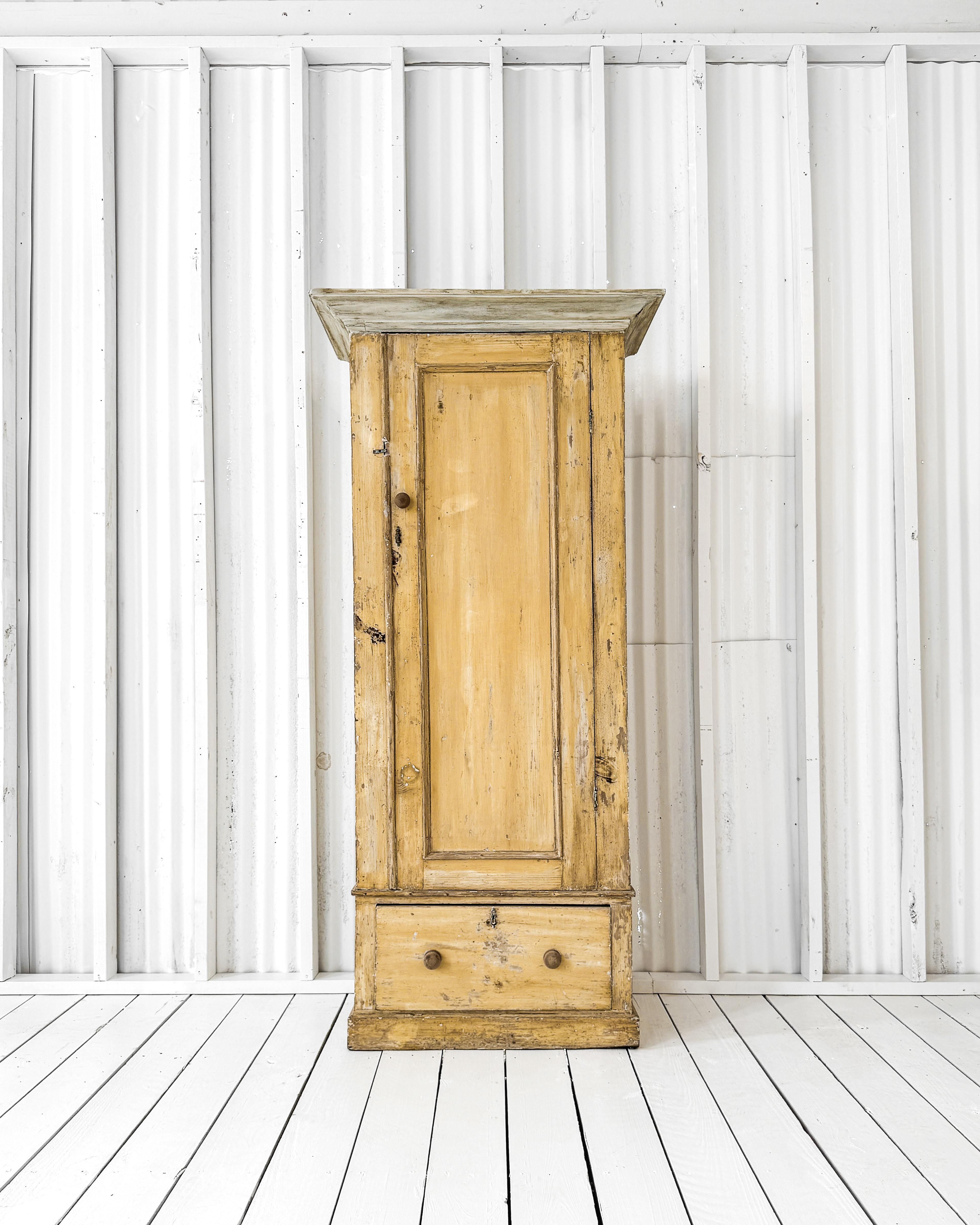 A vintage cupboard featuring a time-worn buttery yellow paint finish. The single molded panel door opens to reveal three shelves. A lower drawer provides additional storage for linens and the like. Decorative trim work and crown molding dress up the