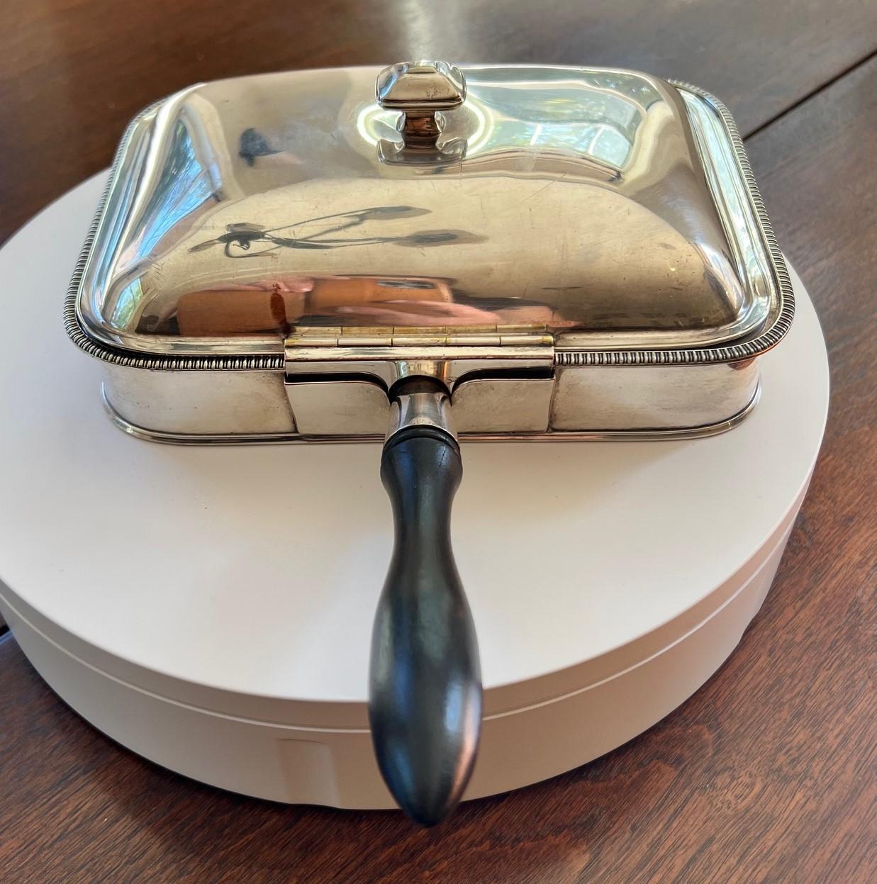 A vintage silver plated English Silent Butler with an ebonised wooden handle. The lid, engraved with a lion, raises to show a rectangular recessed receptacle. The hollow space underneath the recess allows for the safe collection of hot debris such