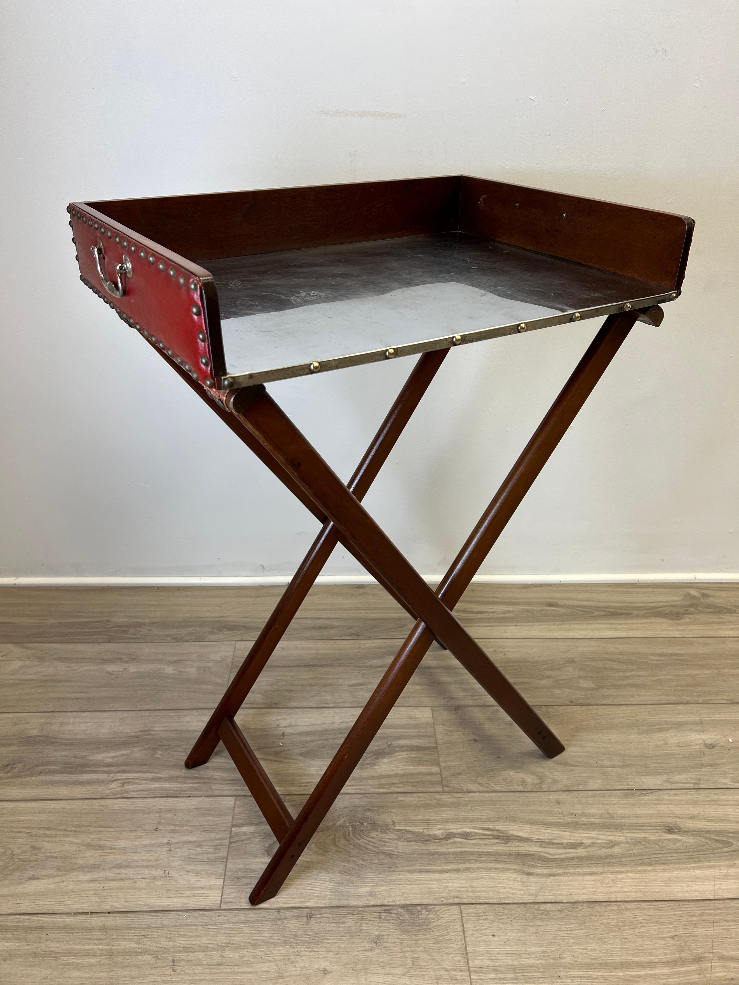 Featured is a wonderful small English vintage bar tray table on a collapsable stand. The stand is made of hinged mahogany legs mounted with a pair of leather straps. The top tray is built of mahogany as well, lined on three sides with red embossed
