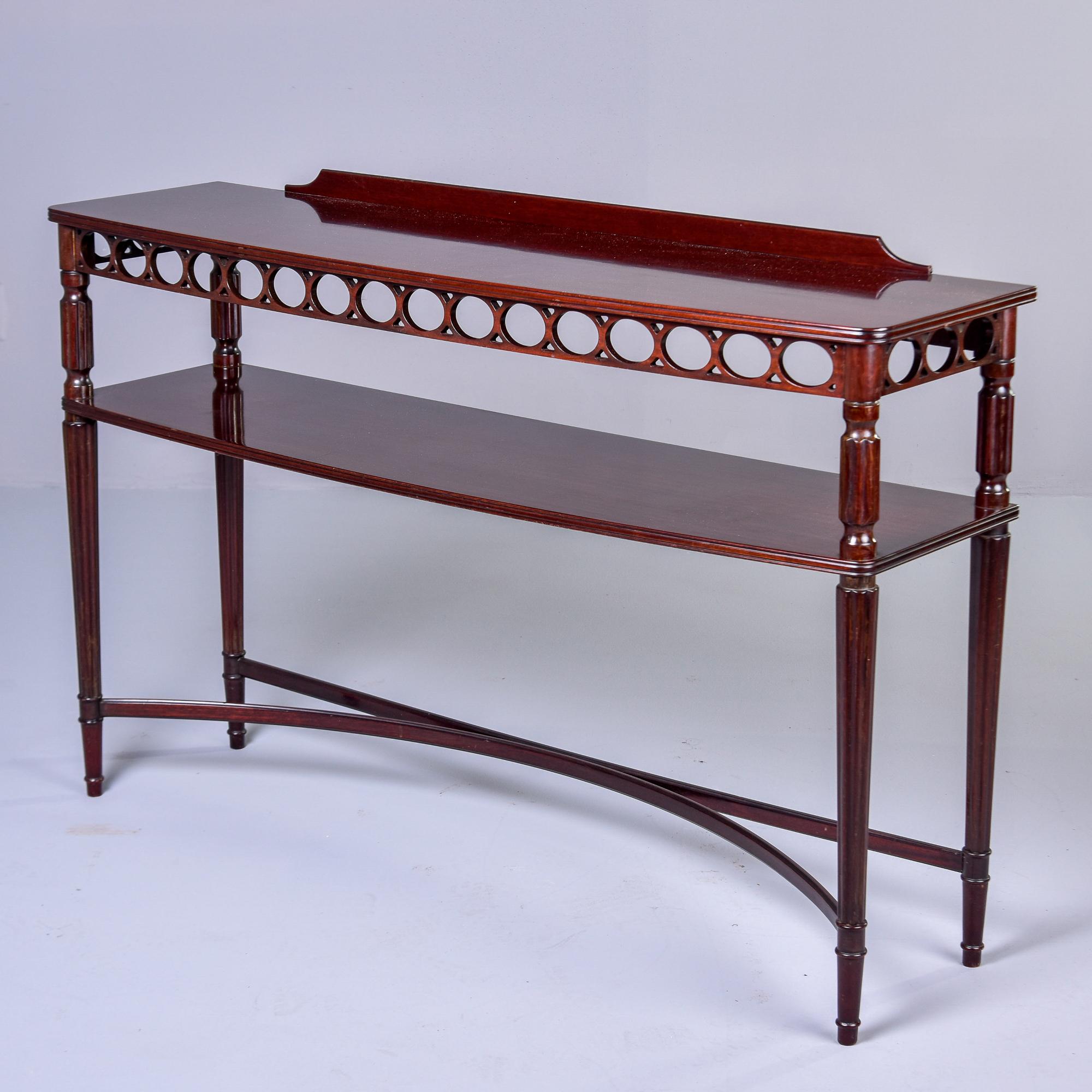 Found in England, this slender two tier mahogany console dates from the 1930s. Table top has a low back panel and an apron with open work, circular design. There is a lower tier and four turned and tapered legs supported by curved X-form stretchers.