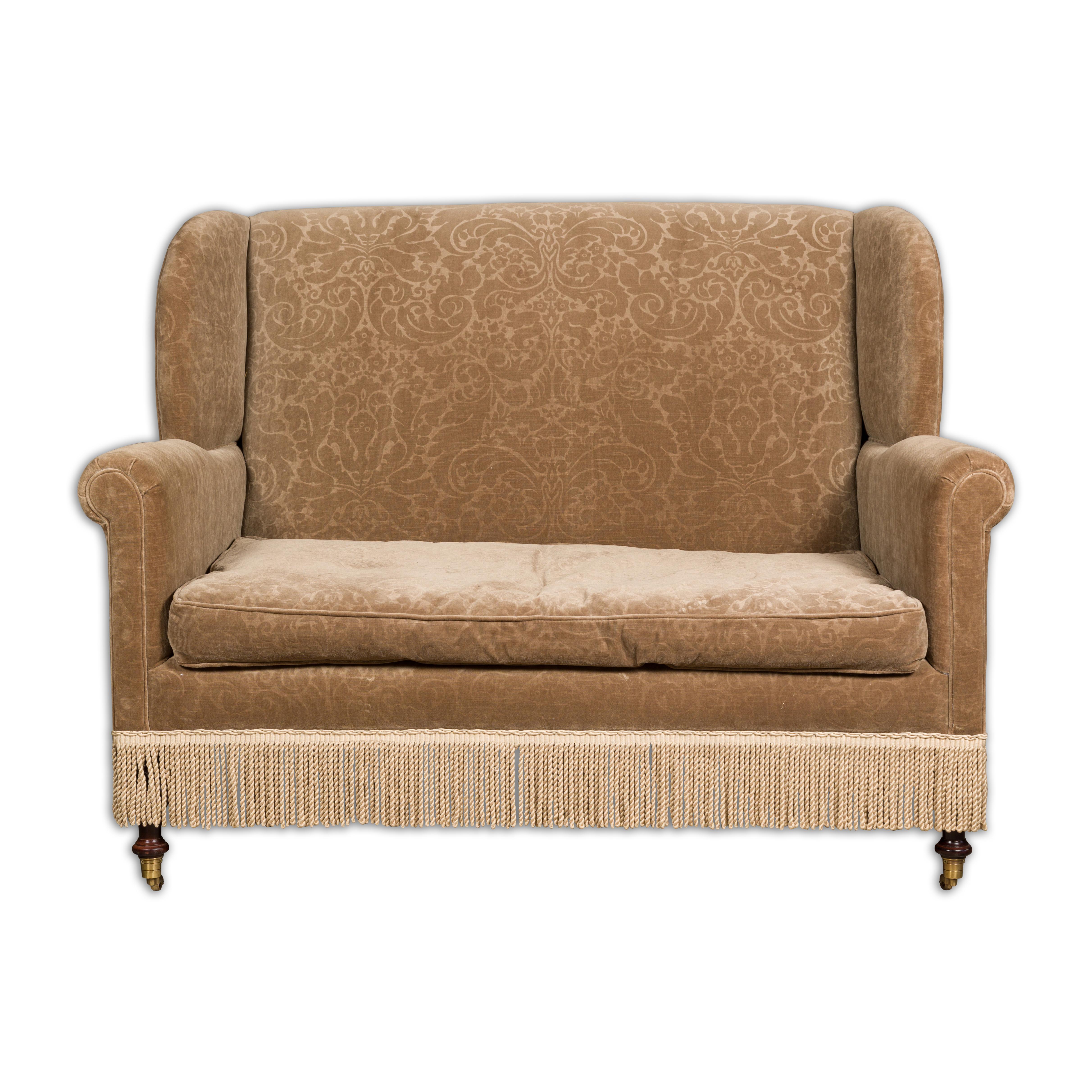 A vintage English wingback loveseat / settee with brown upholstery, out-scrolling arms and casters. This vintage English wingback loveseat, or settee, is a testament to timeless elegance and comfort. Upholstered in a brown fabric adorned with