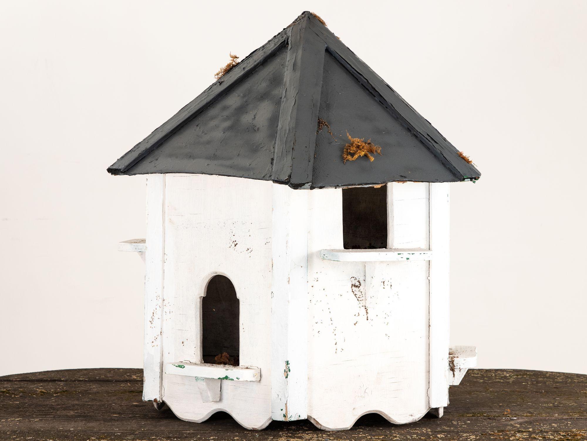 This is a large English country six-sided dovecote from the early 21st c. It is resplendent in weathered white paint, exuding rustic allure. Standing tall, it boasts six pigeonholes of varying heights, creating a staggered visual symphony. Each