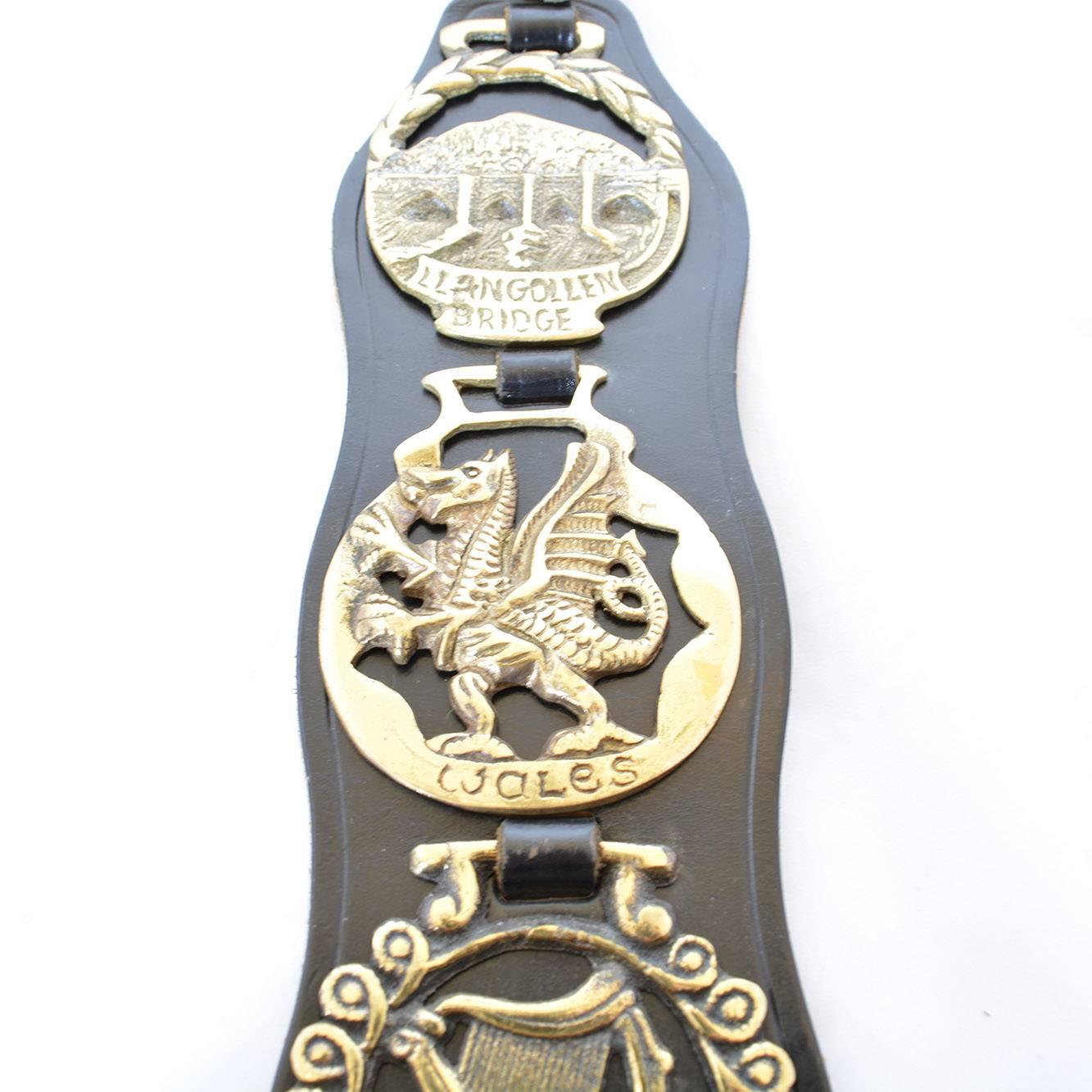 This equestrian themed vintage wall hanging has four horse brasses on a leather strap. The four horse brasses portray various items including a Llangollen Bridge, Welsh dragon, triple harp, and the coracle man. Each object depicted is associated