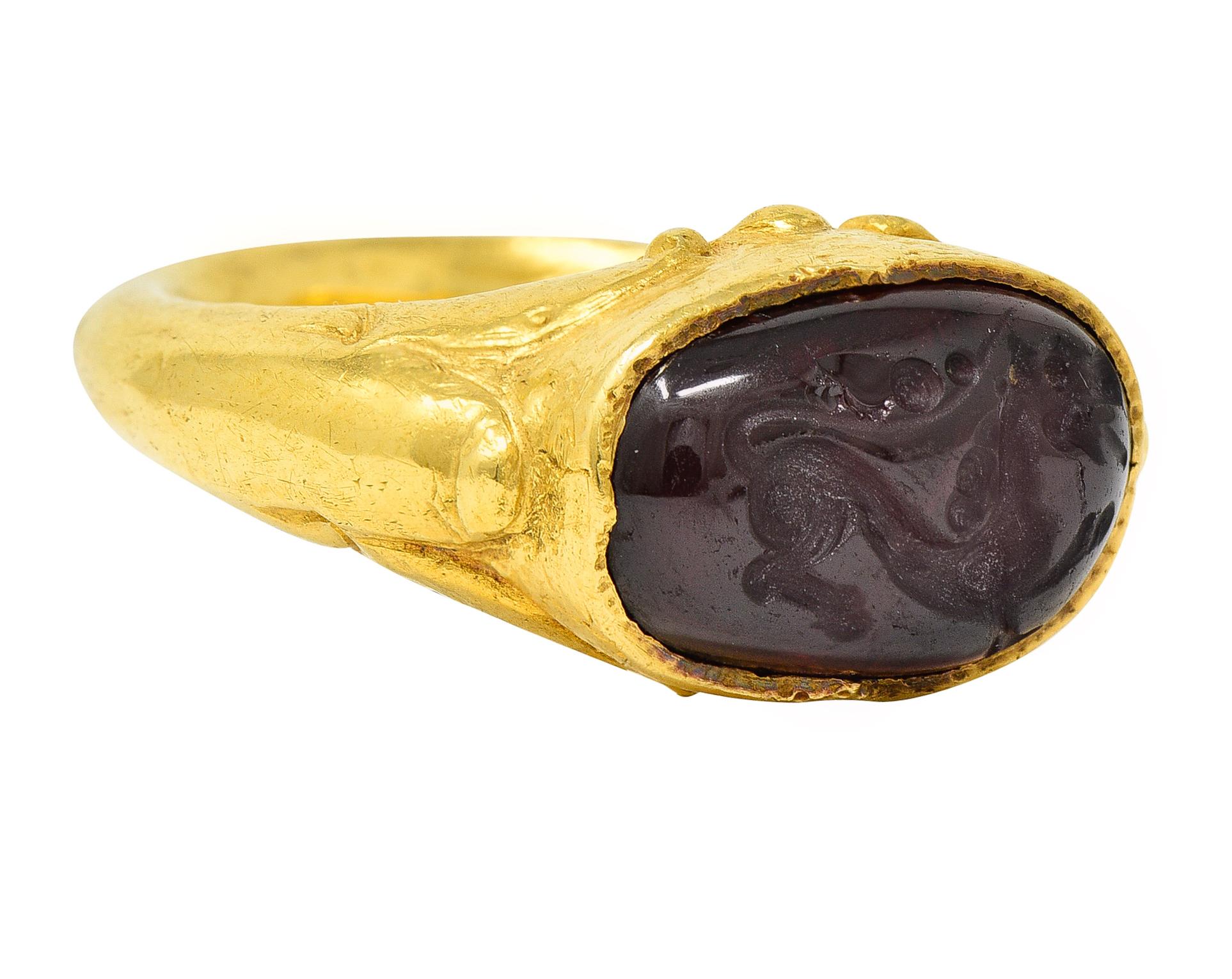 Signet style ring features an oval garnet cabochon measuring approximately 8.7 x 13.0 mm. Dark red in color and deeply engraved to depict a stylized roaring lion. Bezel set East to West in a bombè style mounting. Completed by raised lotus flower