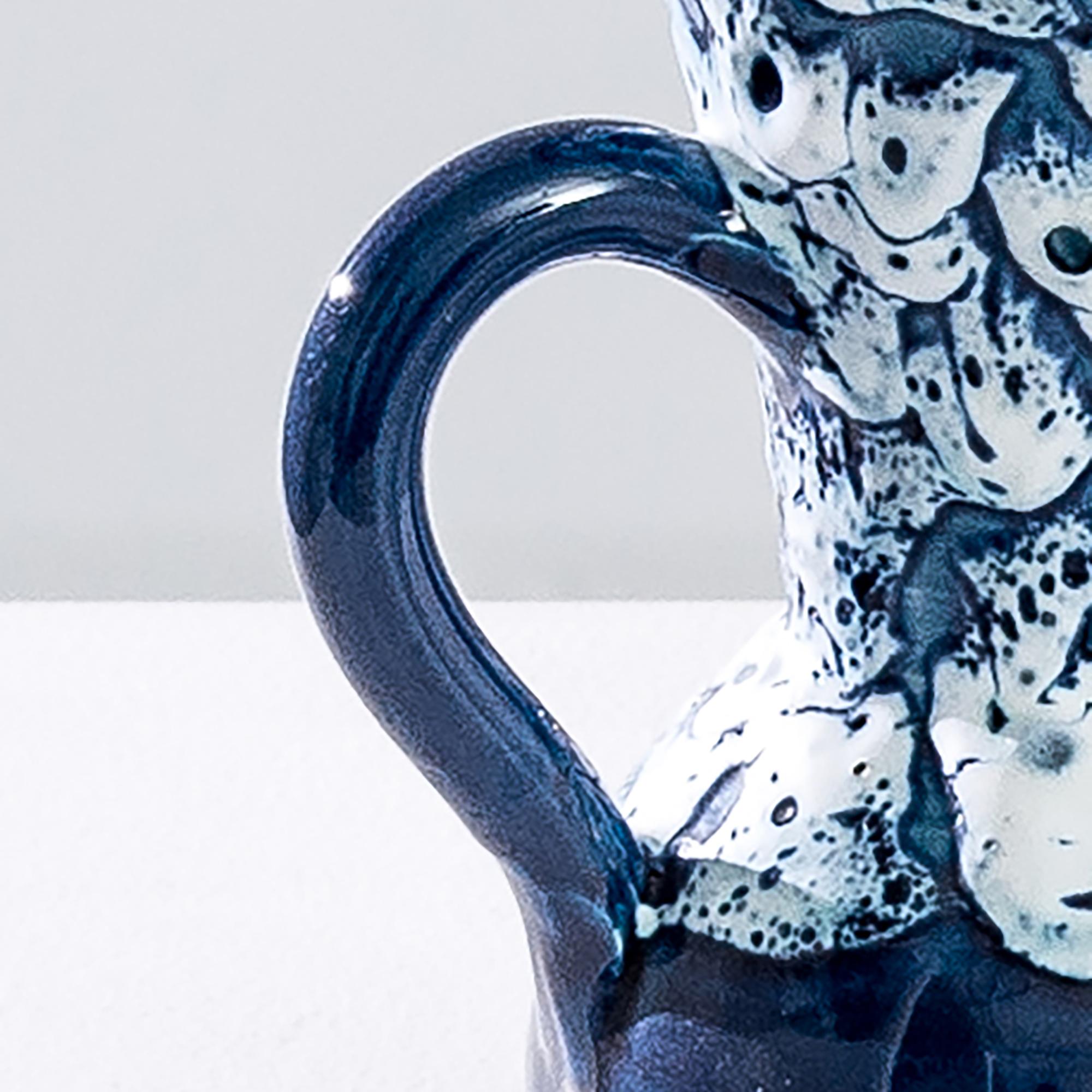 This ceramic vase was produced in Europe in the 20th century. A large neck with two loop handles gives the jug a unique shape, complemented by the round base. The glazed surface is covered with mottled abstract patterns in white and dark blue tones.