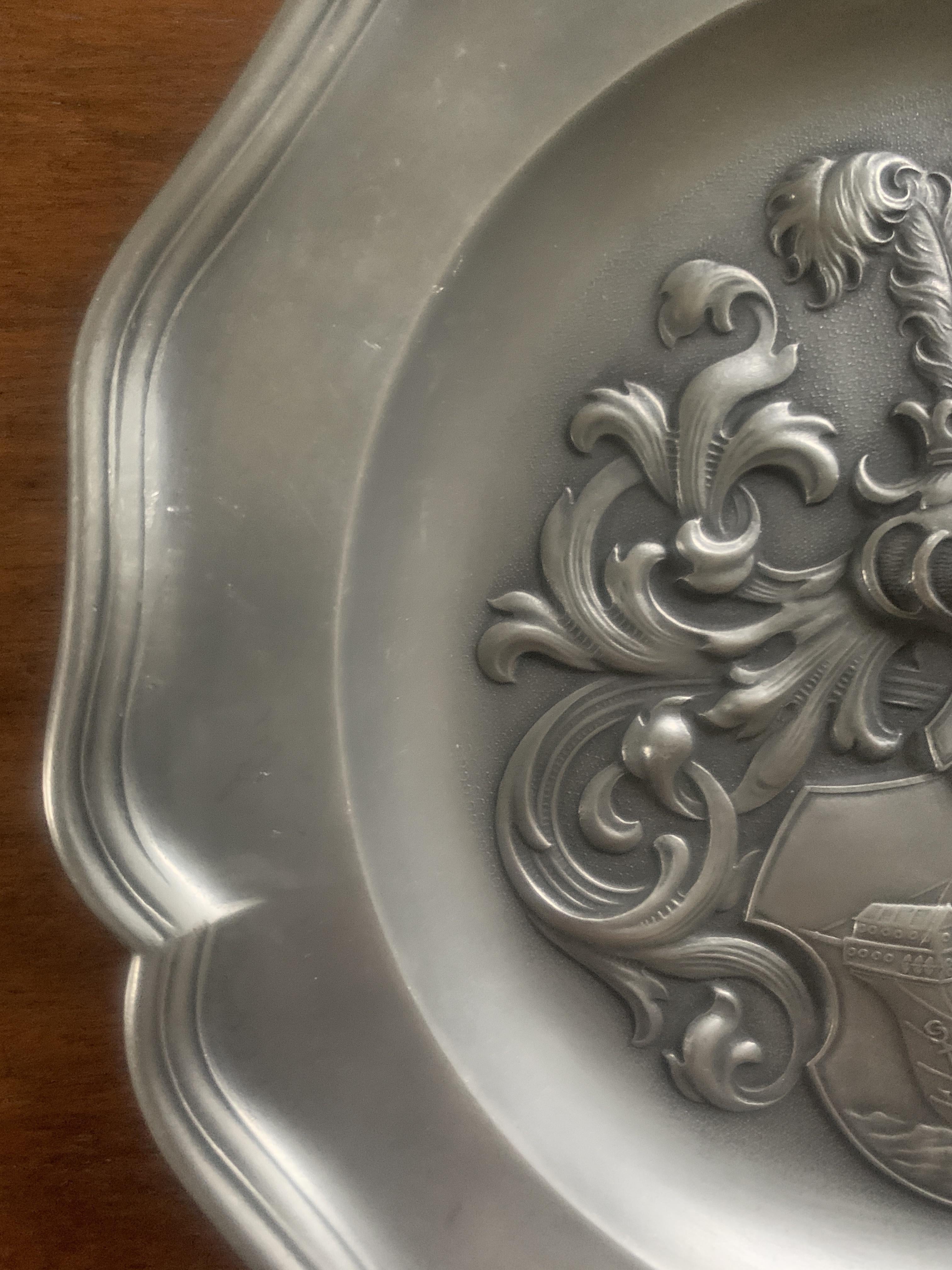A vintage pewter European wall plate with a coat of arms featuring a knight holding a shield with a ship at sea on it.
 