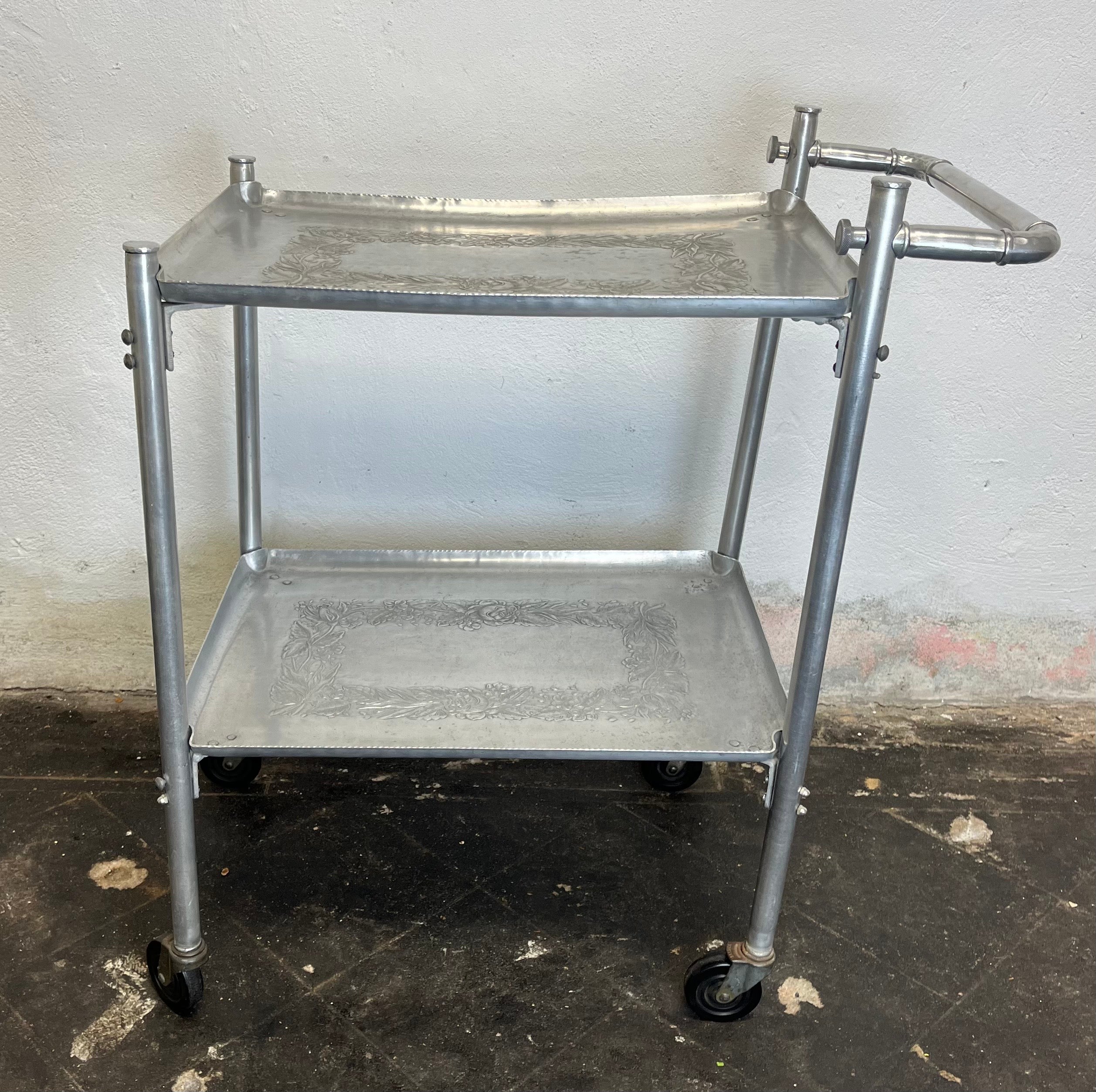 Une belle pièce d'époque. Chariot de service en aluminium martelé à 2 étages. Superbe poignée en faux bambou et plateau au design floral intemporel.
Sera démonté pour l'expédition.