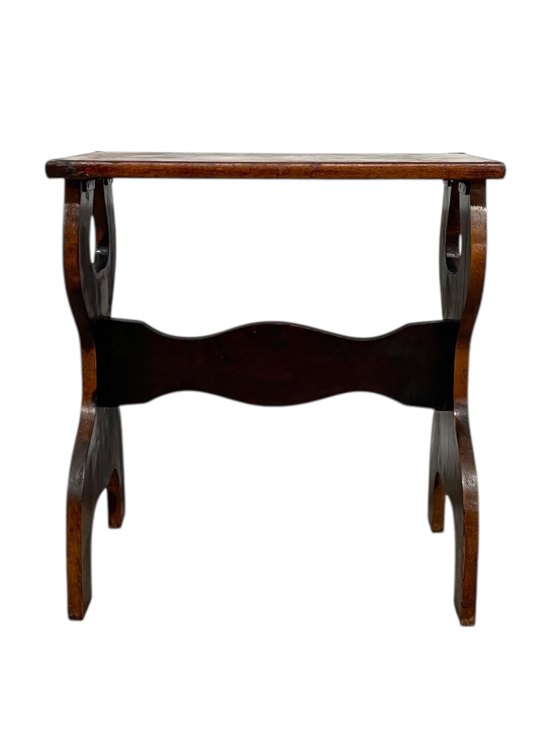 Este soporte para plantas o mesa auxiliar de estilo granjero vintage presenta una silueta rústica con soportes laterales curvados cortados a mano y un sencillo tablero rectangular. La pieza está fabricada en madera maciza con un acabado teñido