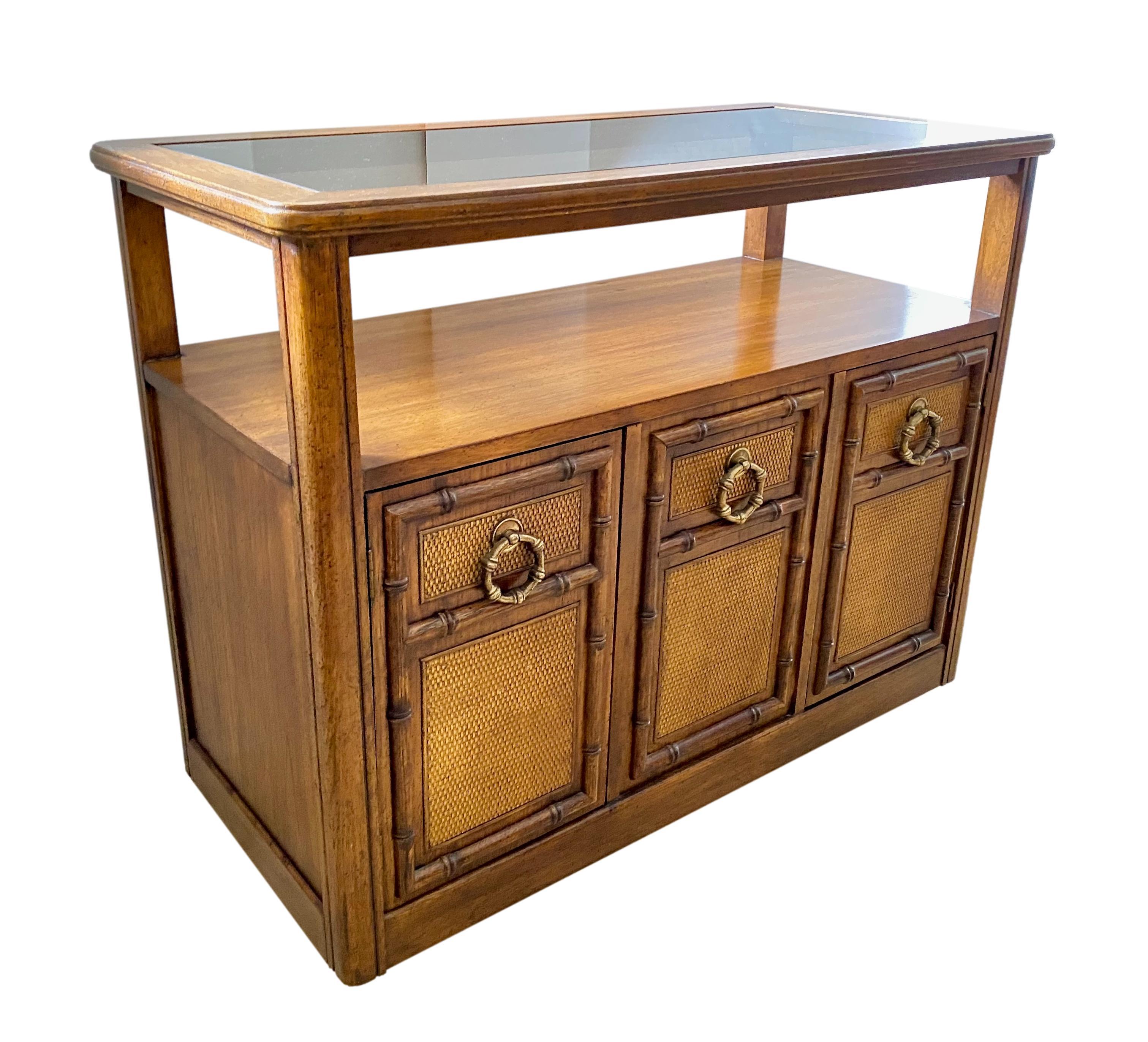 A 1970s Hollywood Regency style console table and two-door storage cabinet on casters, attributed to American of Martinsville. Wood construction with inset smoked glass top, carved faux bamboo accents, woven reed paneled cabinet doors and bamboo