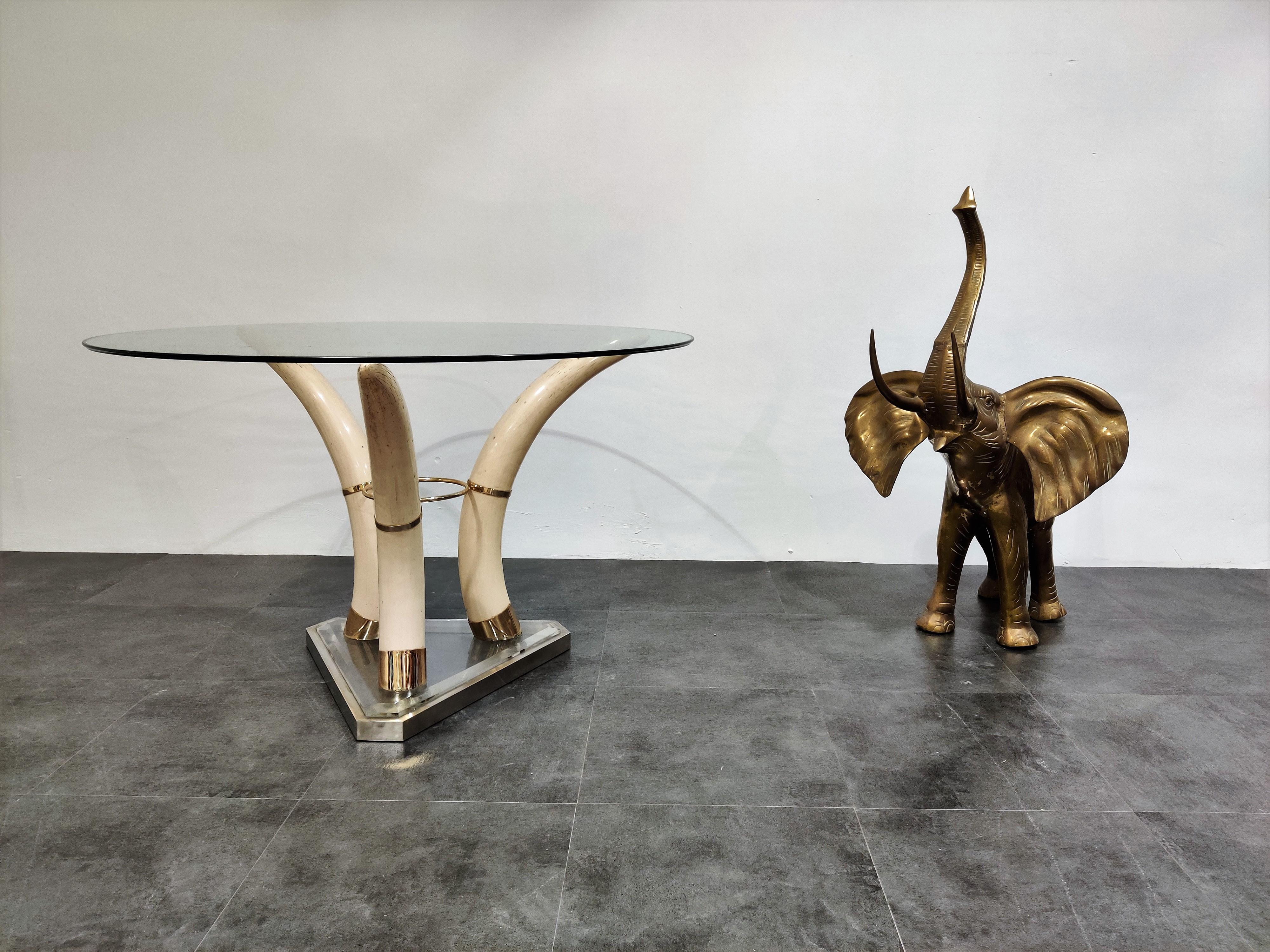 Elegant center table featuring faux elephant tusks made from resin mounted on a metal and lucite base with a round beveled glass top.

The tusks are held upright by brass cups and fastened with a brass center ring. 

Real eye catching occasional