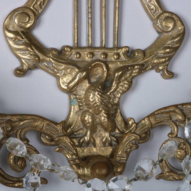 A vintage pair of figural wall sconces offer cast brass construction in lyre form with central phoenix having two foliate form arms terminating in candle sockets, hanging cut crystal highlights throughout, circa 1930.

Measures: 12