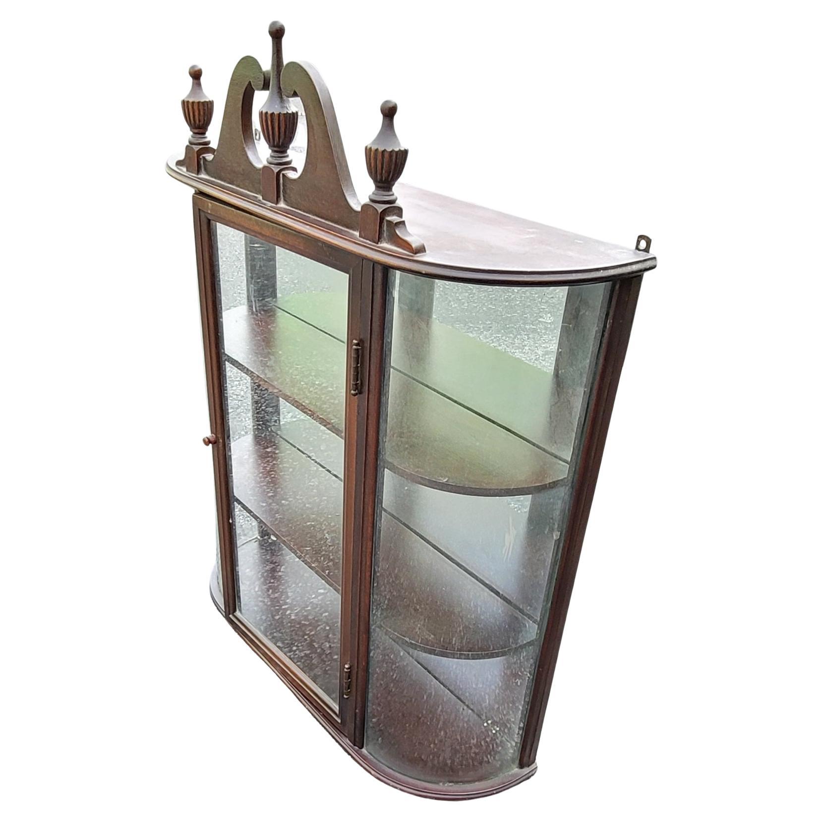 Une vitrine murale vintage de style Feder avec une façade en verre arrondi, un intérieur en miroir et trois étagères, surmontée d'un fronton à volutes et de trois épis de faîtage en forme de trophée, produite par Ferguson Brothers Manufacturing