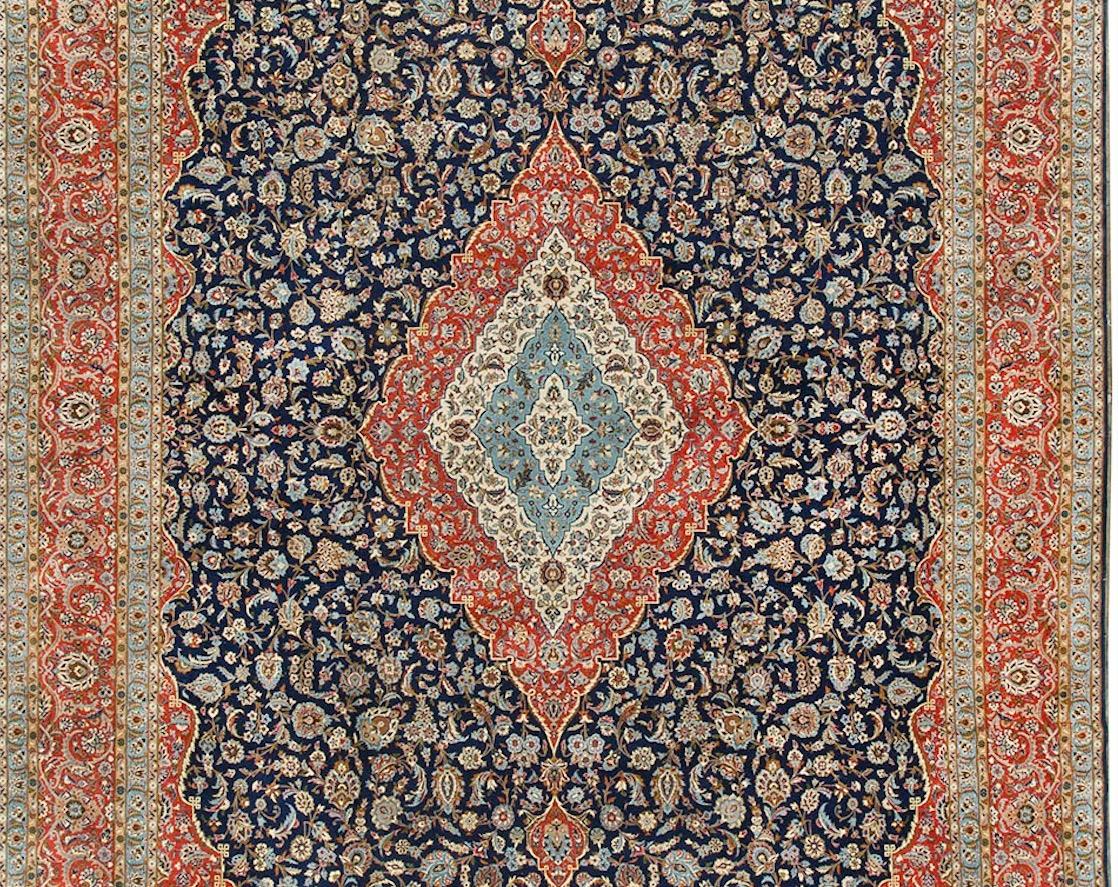 The central motif, surrounded by a magnificent field of flower heads, combined with the exquisite use of colors all on a dark navy ground give this vintage Kashan rug a total effect of strength coupled with a dramatic feel. Measures: 13'2