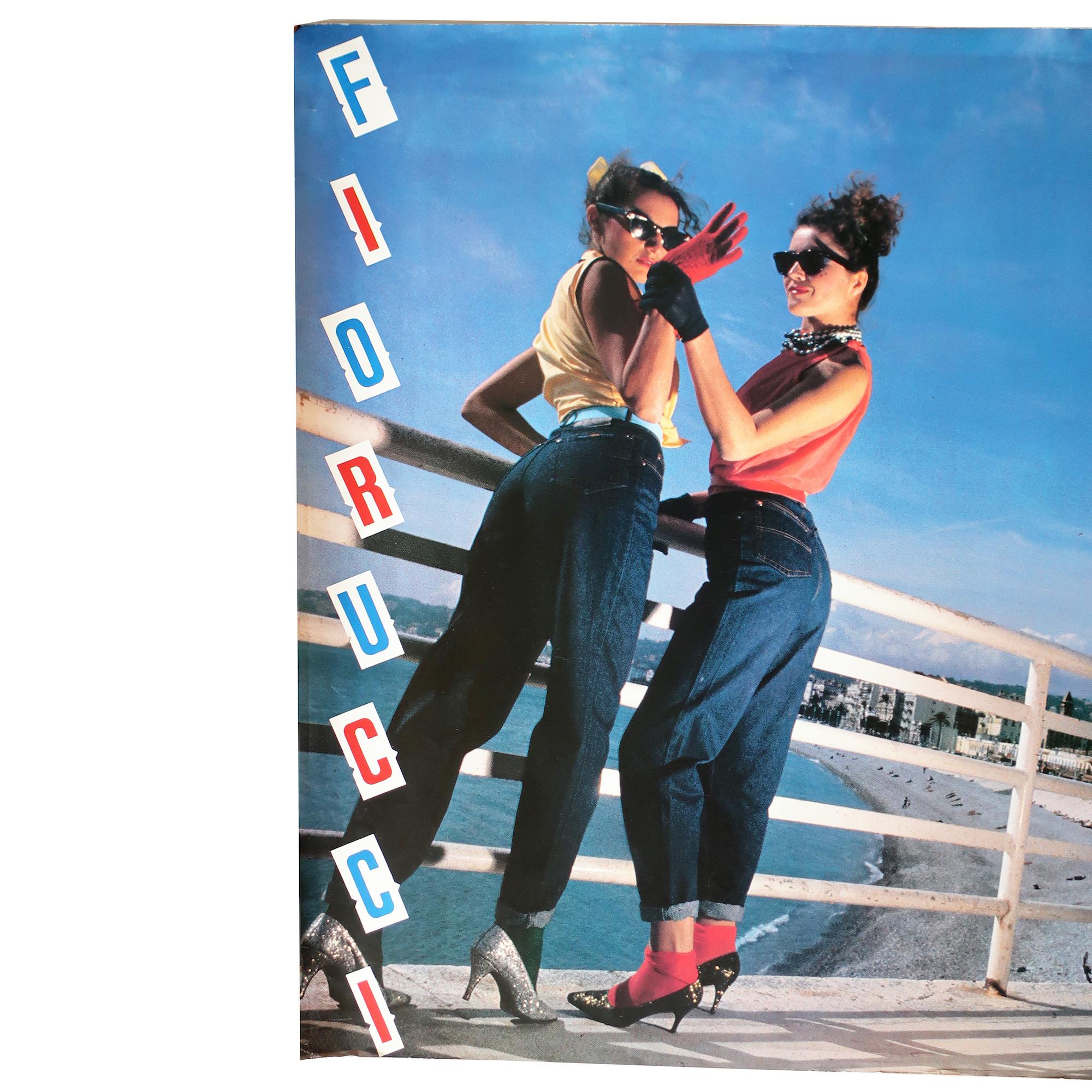 A vintage and rare Fiorucci poster with two women in high heels and jeans waving while a motorcyclist pulls up behind them on a pier by the sea.  The photo is beautifully lit and the poster includes slightly diagonal FIORRUCI lettering on the left