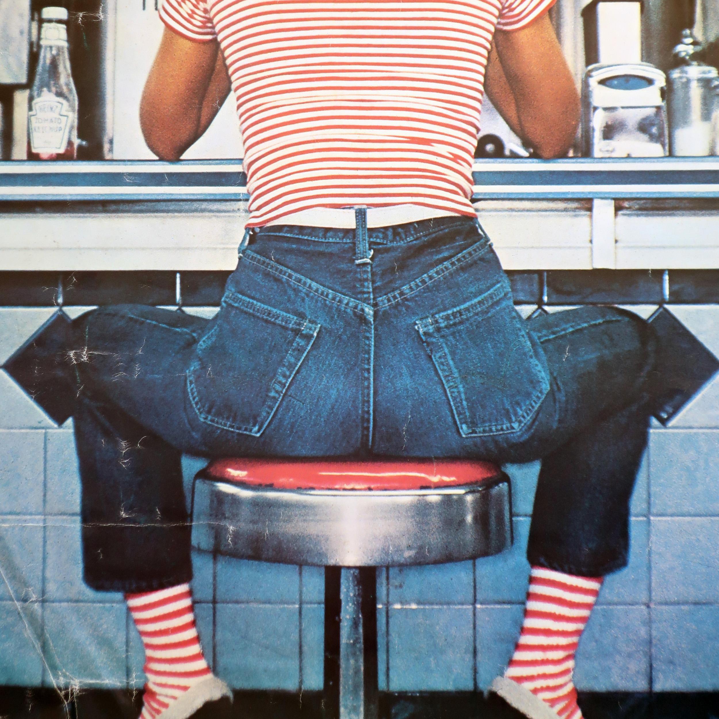 A vintage and rare Fiorucci poster featuring a woman sitting on a stool at a diner counter wearing an iconic Fiorucci striped shirt, matching striped socks, and blue jeans. Photograph is by Oliviero Toscani, the great Italian fashion photographer