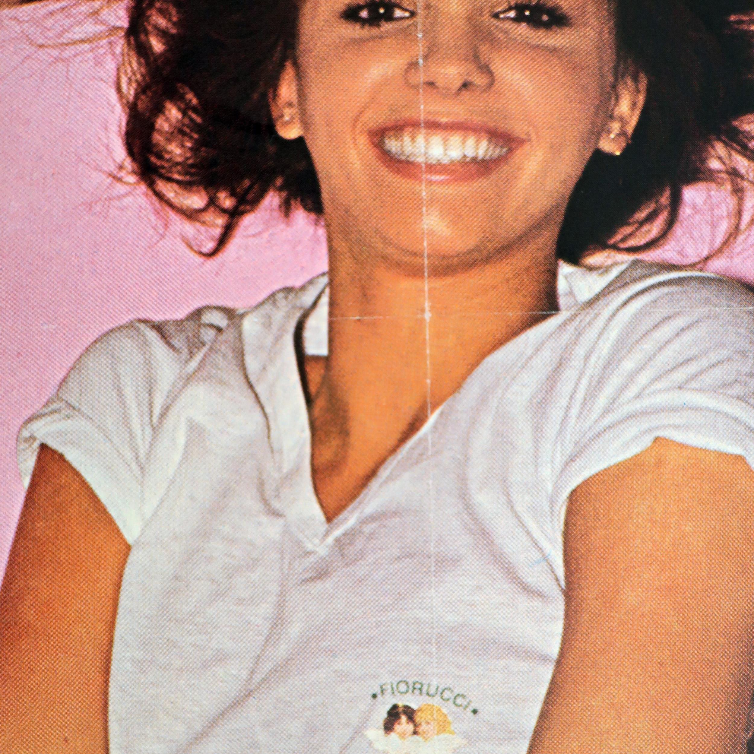 A vintage and rare Fiorucci poster featuring a beautiful pants-less woman caught in mid-disrobing of a white v-neck t-shirt with the iconic Fiorucci two angels logo. Model poses in front of a pink background and Fiorucci branding (“Fiorucci Sweet