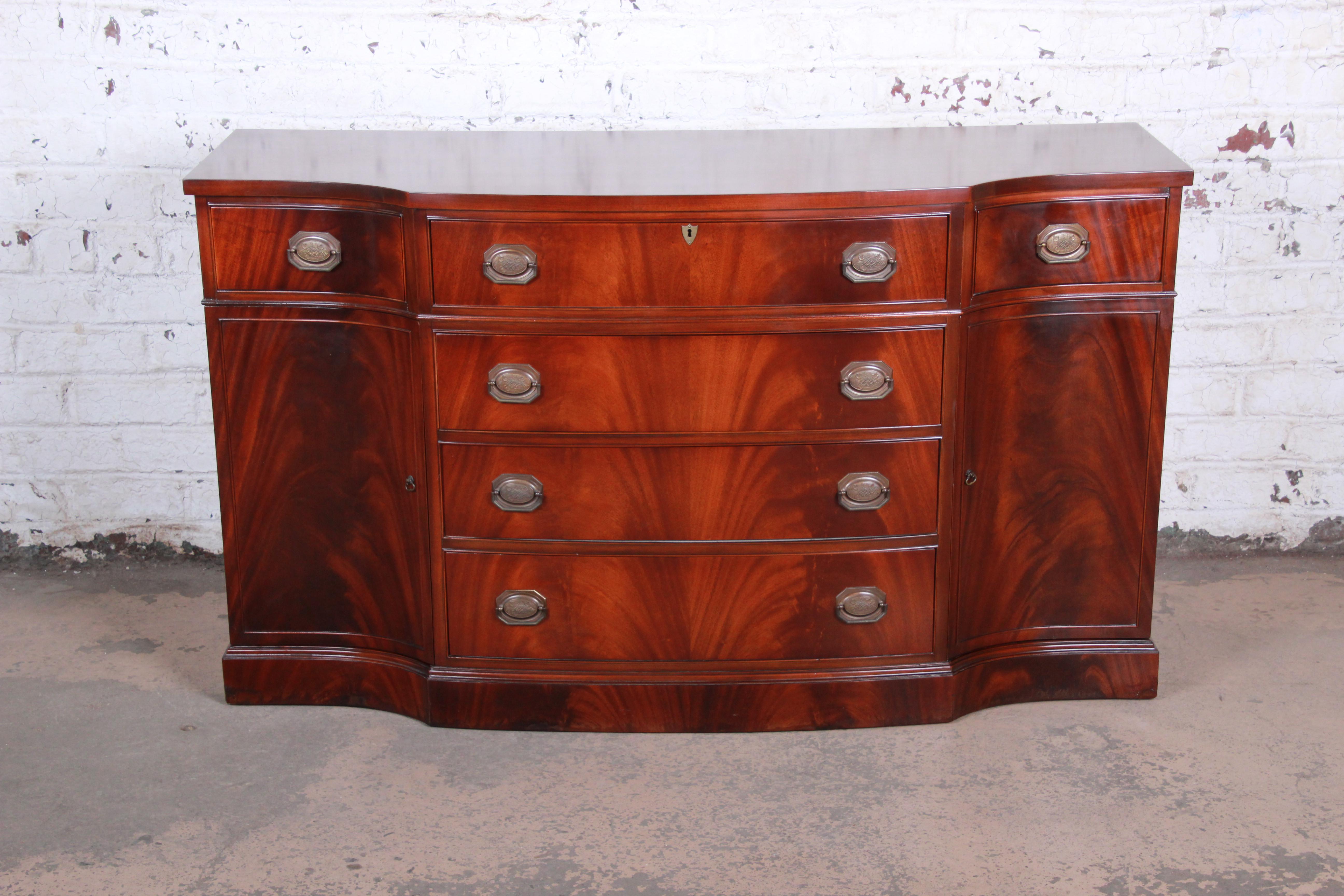 A gorgeous mahogany bow front sideboard or buffet server, circa 1940s. The sideboard features stunning flame mahogany wood grain with a bow Front Design and original brass hardware. It offers ample storage, with six dovetailed drawers and shelved