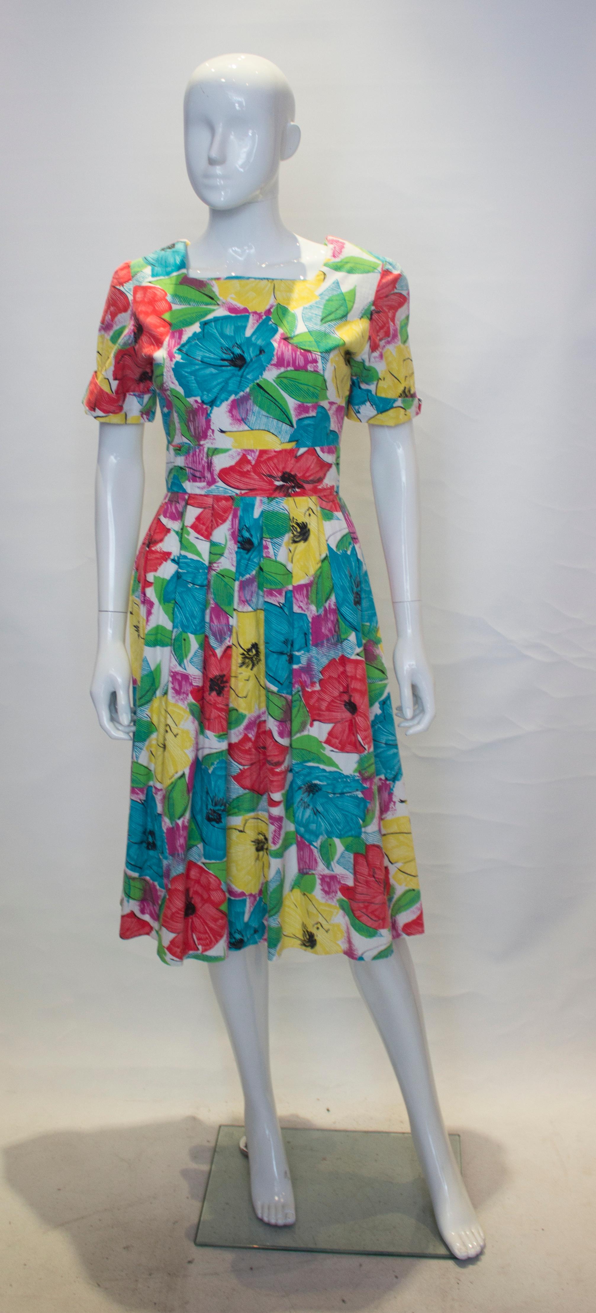 A headturning dress for Summer. This flora cotton dress in an attractive mix of  green , yellow and pink, has a square neckline and backline with band detail at the back.  It has short sleaves with turn back cuffs, pleats at the waist and a button
