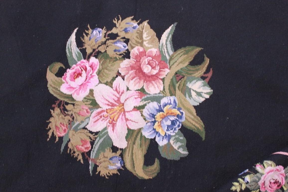 This large-scale vintage needlepoint rug / tapestry features a striking black ground, punctuated by alternating large and small bouquets of multicolored flowers. The floral motifs are surrounded by an elegant floral border, enhancing its rich,