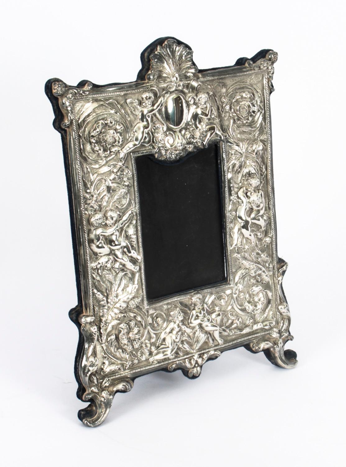 A truly superb large decorative sterling silver photo frame by Neil Lasher Silverware with hall marks for London 1995.

The frame features beautiful chased and embossed decoration with flowers, foliage and cherubs in high relief and a navy velvet