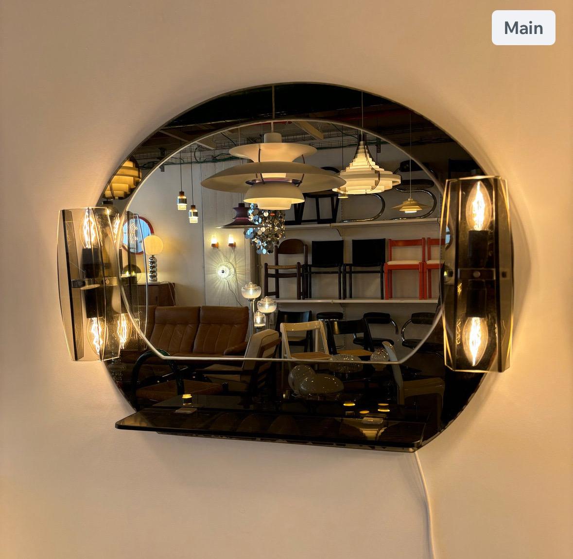 Ce miroir vintage Fontana Arte, conçu par Luigi Fontana dans les années 1970 en Italie, est un exemple frappant du design moderne du milieu du siècle. Fabriquée en verre miroir, cette pièce présente un miroir central flanqué d'une paire de douilles