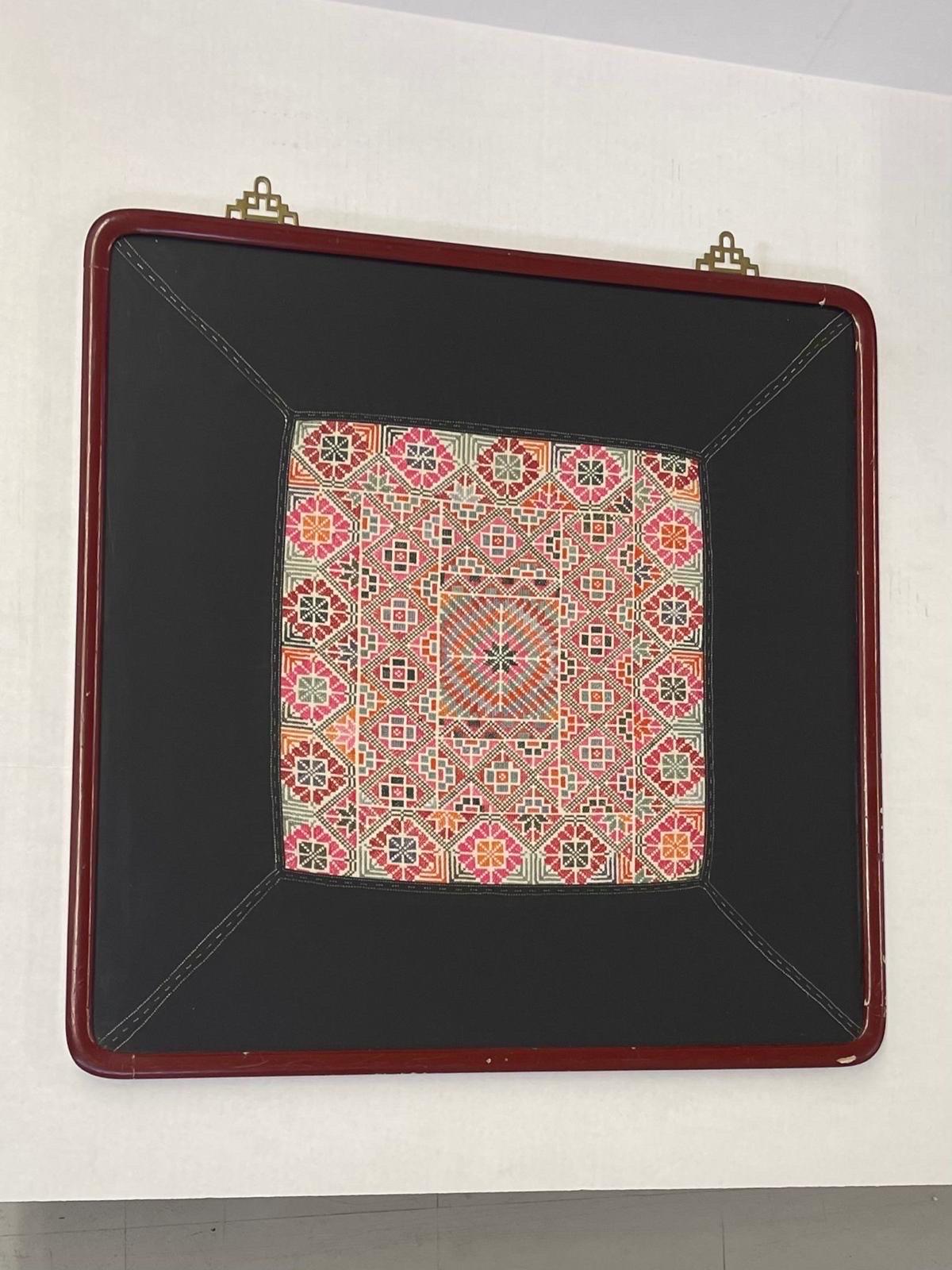 This Piece features two brass toned hinge hooks coming from the top of the frame. Maroon colored frame. Embroidered Textile may be of Chinese origin based on Research. The Textile is stitched on to a black fabric acting as the border. Vintage