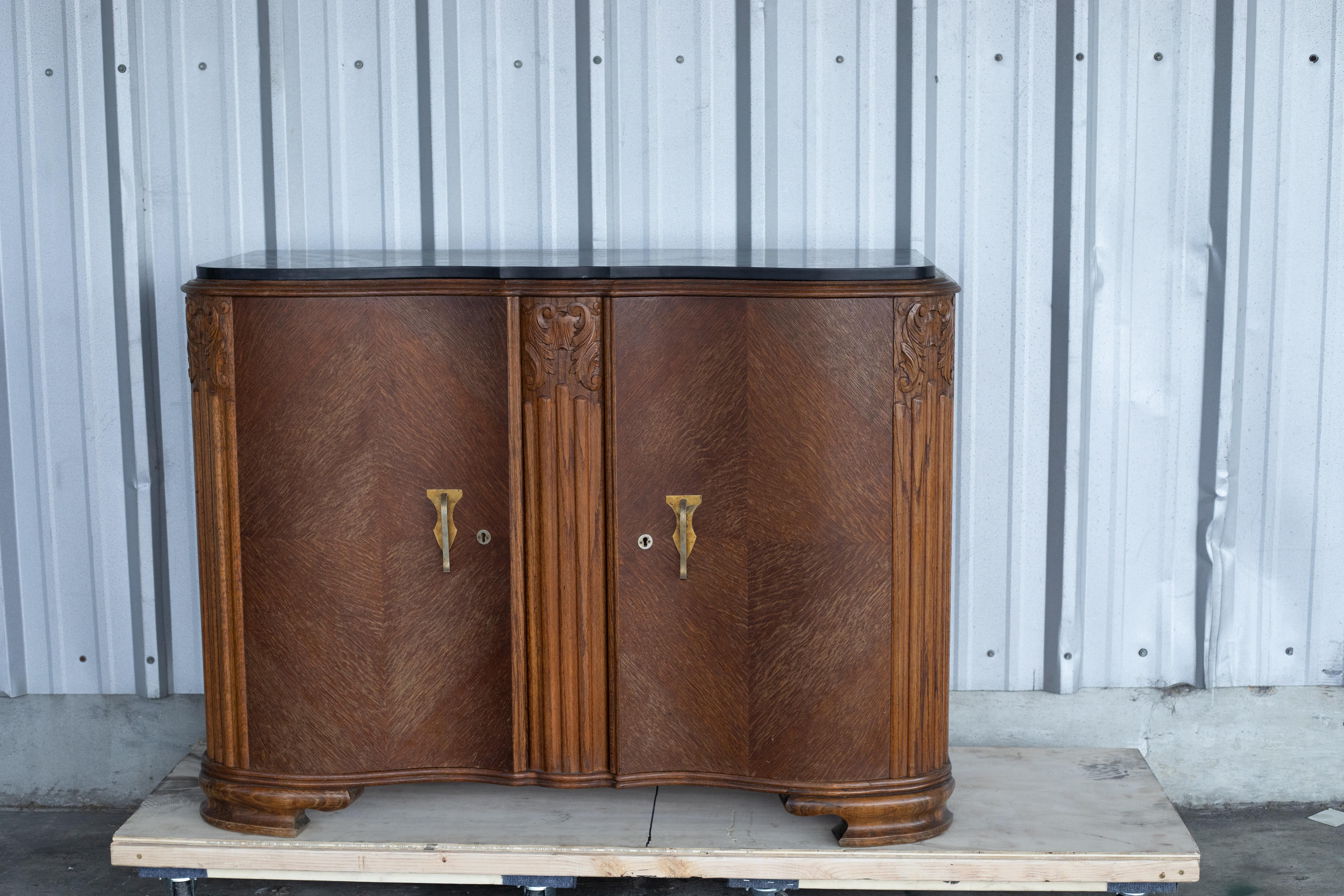 Elegante aparador Art Déco francés, de los años 30-40. Esta pieza presenta un frente suavemente curvado con un llamativo chapado de roble con motivos chevron, pilastras estriadas talladas con motivos florales y atrevidos herrajes de latón. La parte