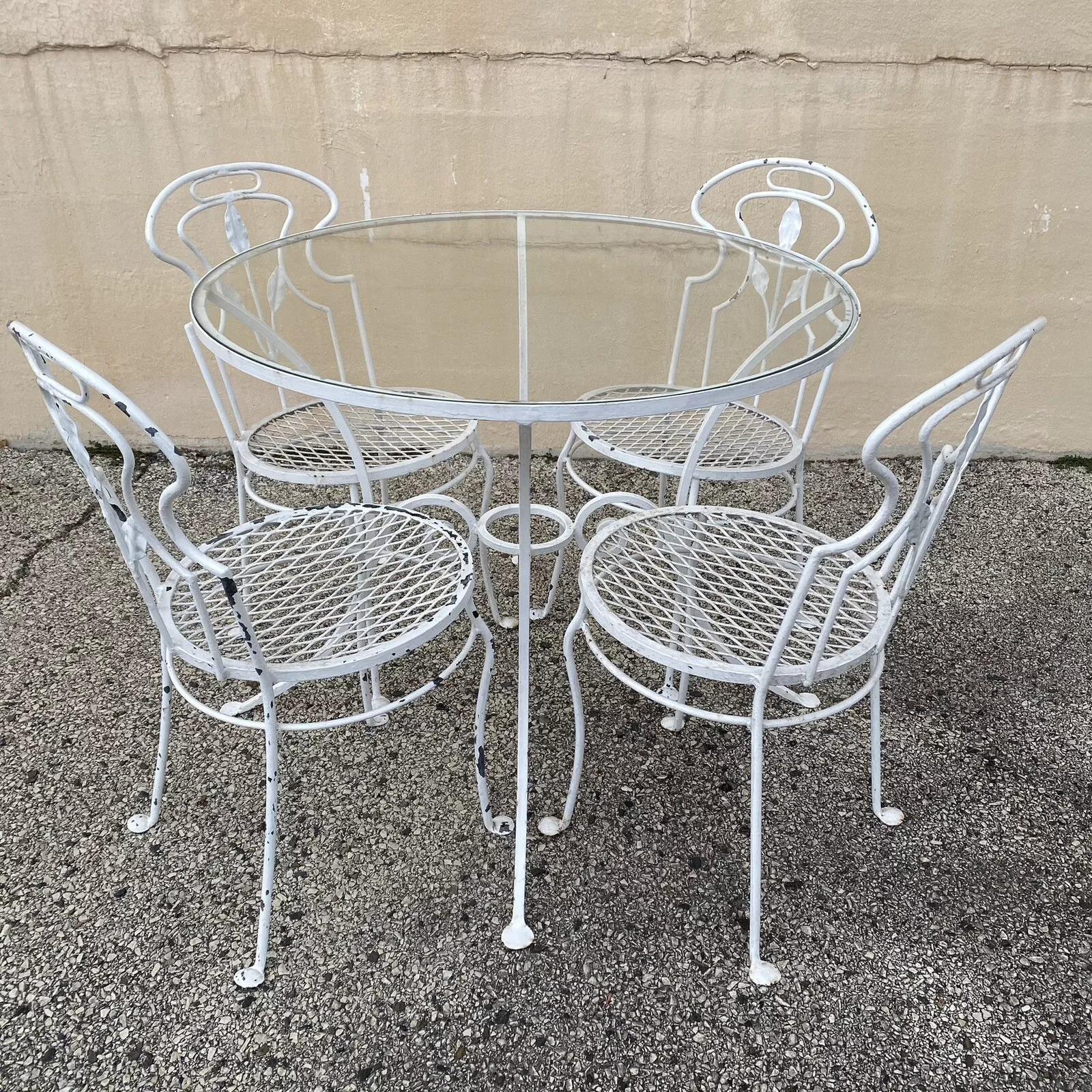 Vintage French Art of Vintage Style Wrought Iron Garden Patio Dining Set - 5 Pc Set. L'ensemble comprend 4 chaises de salle à manger dont le dossier est orné de feuilles et dont l'assise est ronde et perforée. La table comprend un plateau rond en