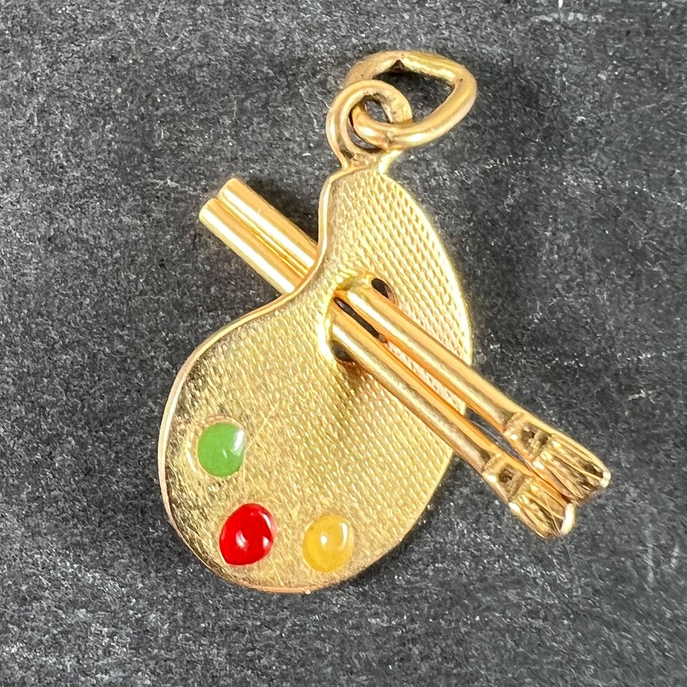A vintage 18 karat (18K) yellow gold charm pendant designed as an artist's palette with three blobs of paint represented in green, yellow and red enamel; with a pair of paint brushes through the thumb hole. Stamped with the eagle's head mark for 18