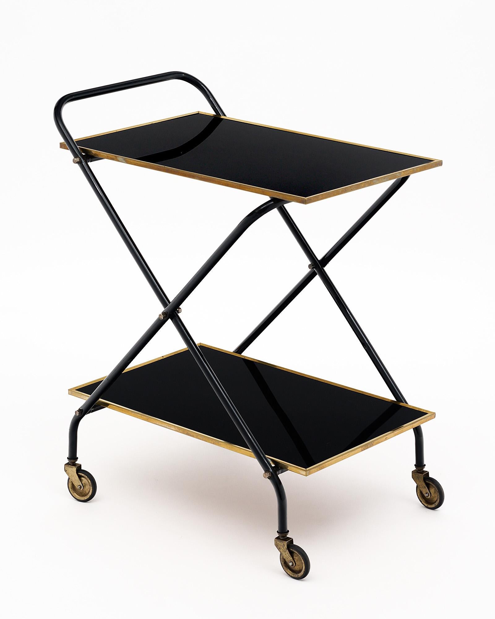 Vintage bar cart from France made with two shelves of black glass. Each shelf is trimmed with brass and the piece is supported by four original brass casters. The structure is black lacquered metal in a curule x shape.
