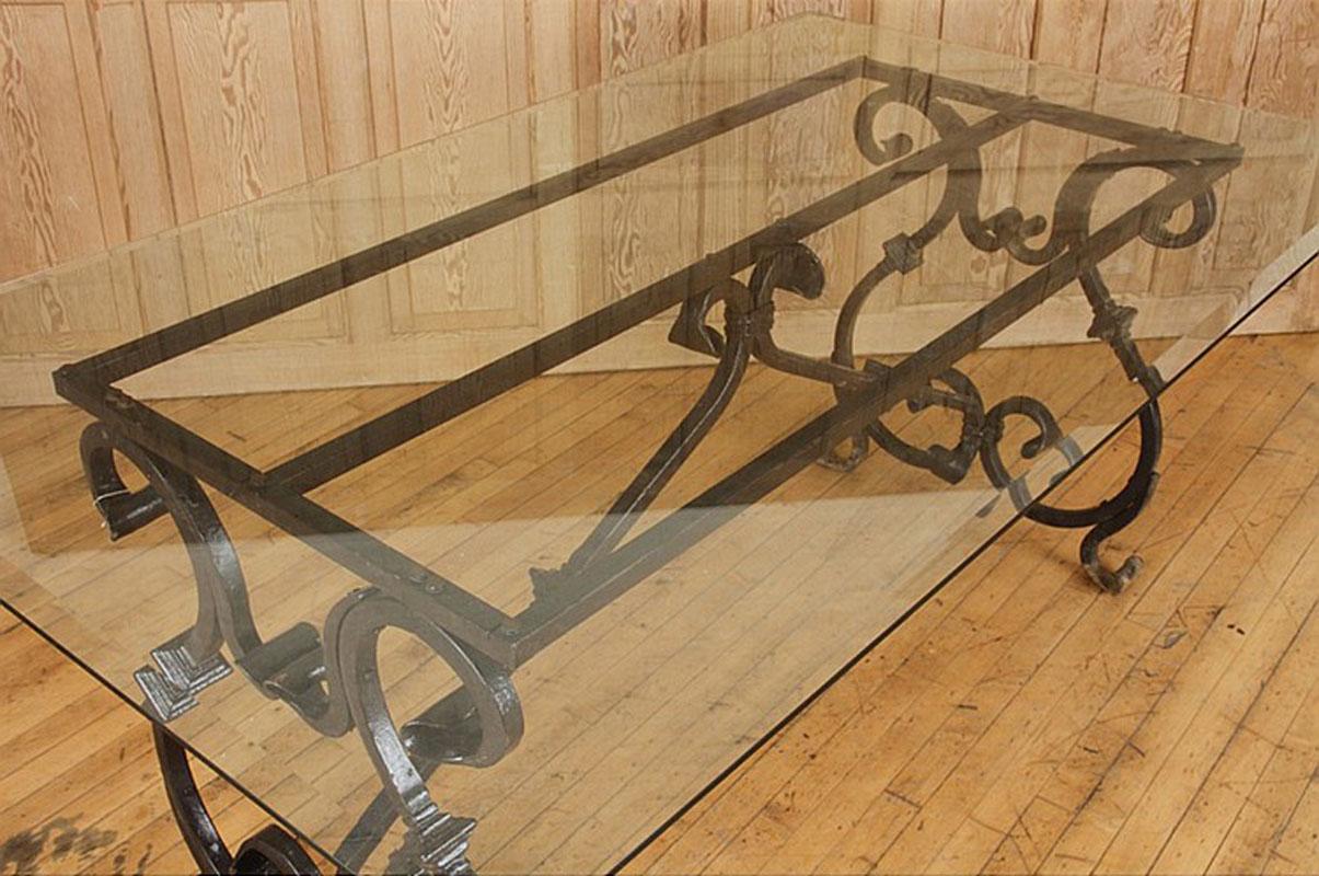 A French Baroque style hand-forged iron table base with glass top.  The trestle base features two lyre-shaped legs adorned with a weathered rust finish, connected to the underside of the top with a curved stretcher. The delicate volutes, joined to