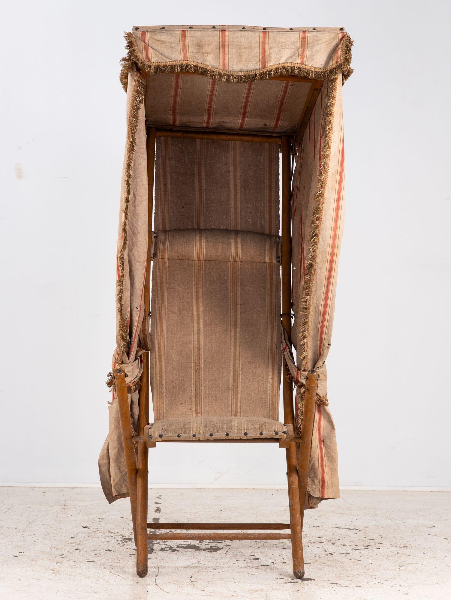This French folding beach chair features a charming built-in canopy, crafted from striped and fringed canvas in warm, weathered tones. The wood frame folds neatly, while the draped fabric enclosure offers both sun protection and a sense of vintage