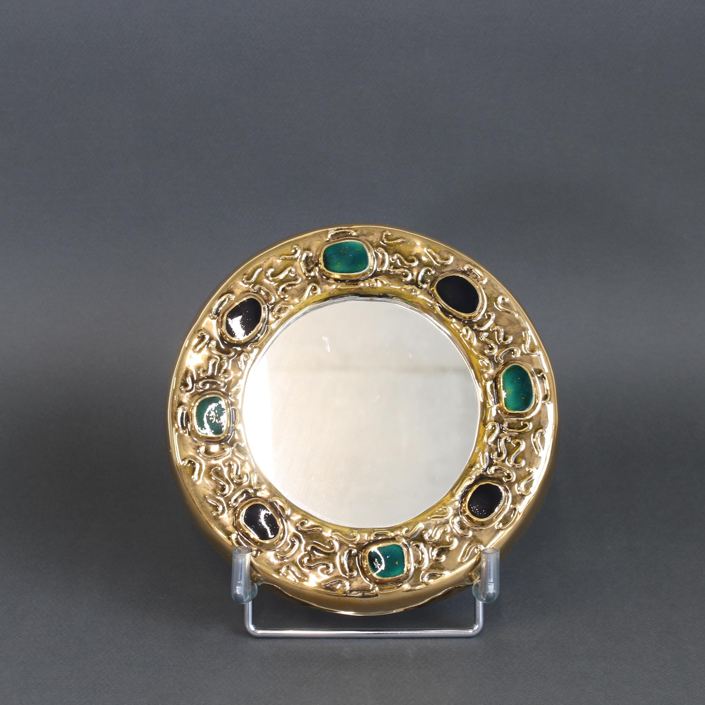 Miroir mural décoratif en céramique dorée de François Lembo (circa 1960s-70s). Cadre en céramique en forme de hublot agrémenté de cabochons émaillés colorés en vert et noir, imitant les pierres naturelles. La pièce est dans l'ensemble en très bon