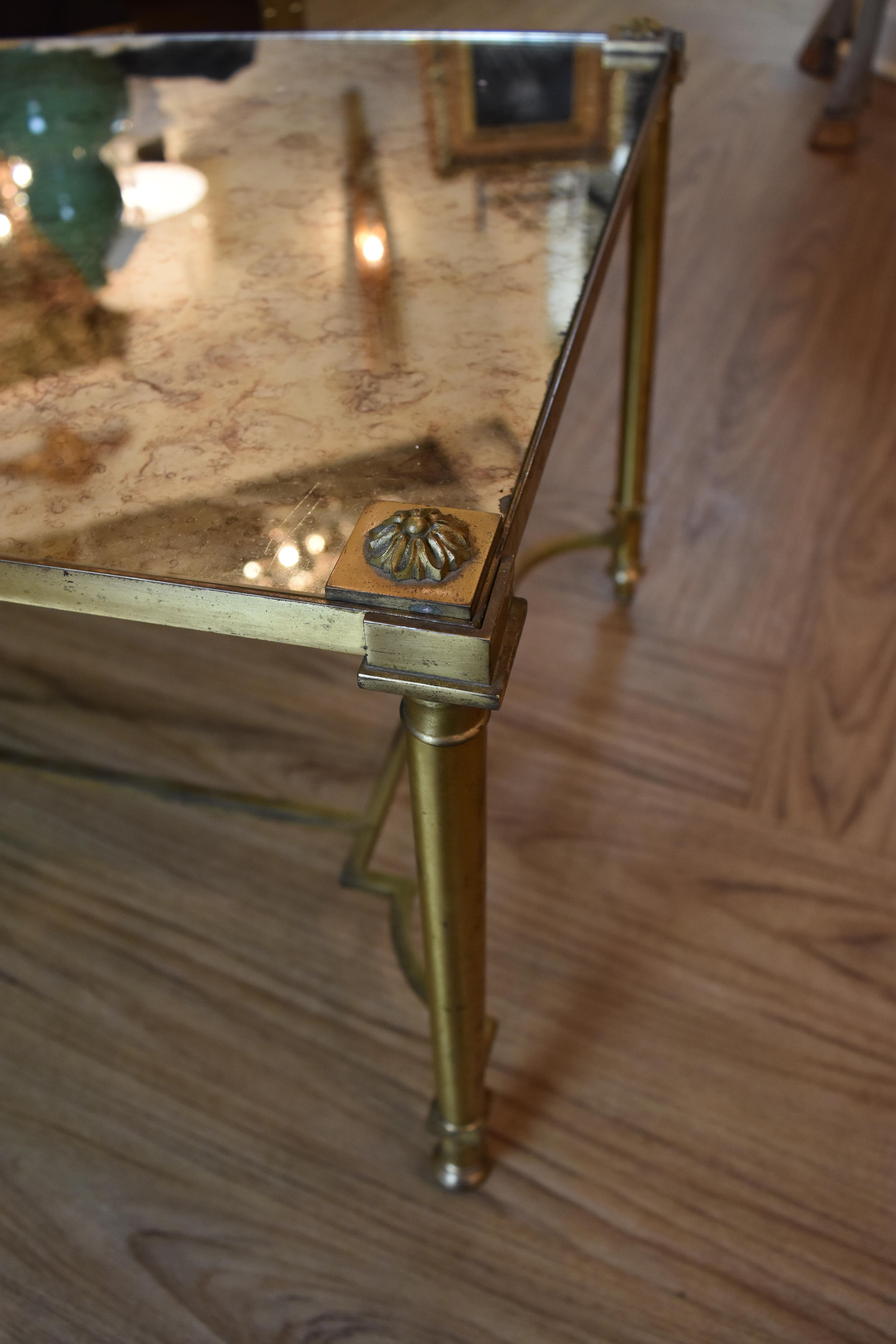 This vintage French coffee table features a brass base and a lovely antiqued mirrored top. Each top corner is inset with a brass rosette on a brass plate. The simple neoclassic design of this coffee table would mix well with both traditional and