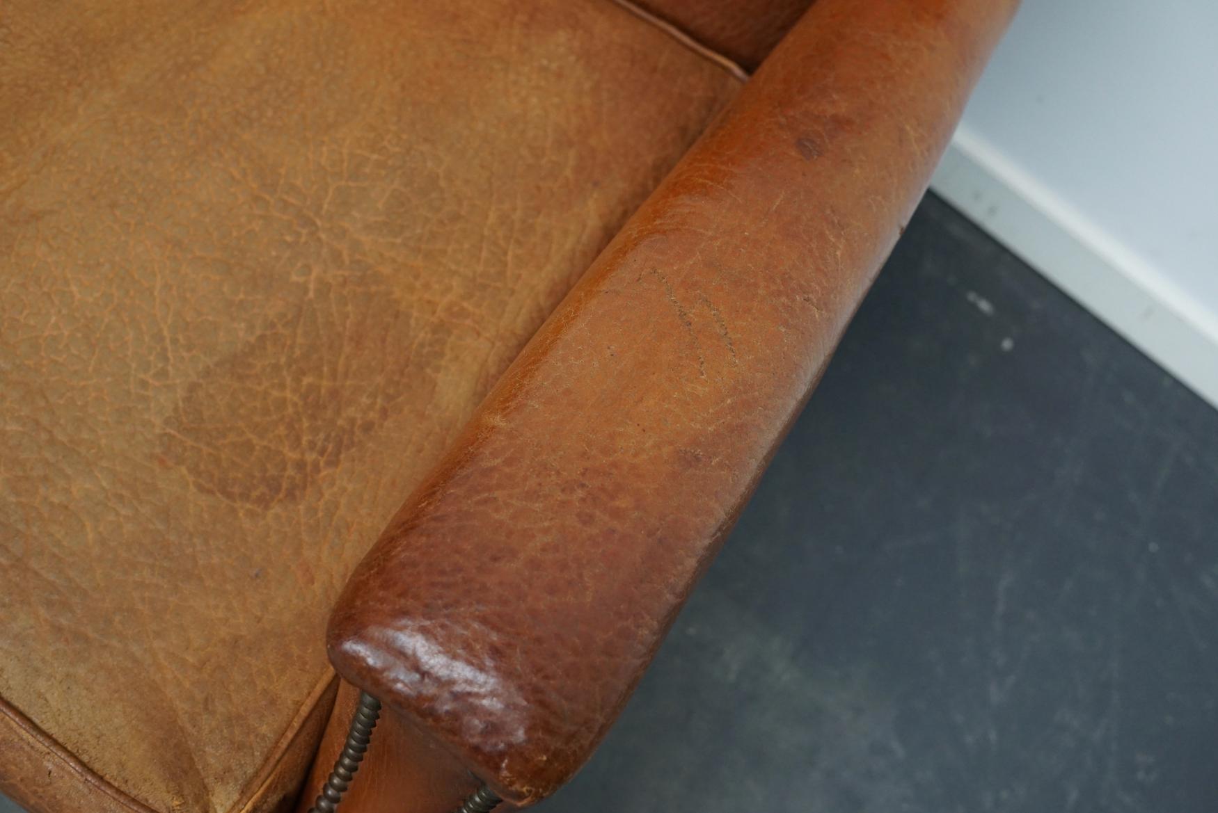 This leather club chair comes from France and was made circa 1940s. It is upholstered with cognac-colored leather and features metal rivets and wooden legs. It remains in a well-used vintage condition with signs of use and age but the chair is still