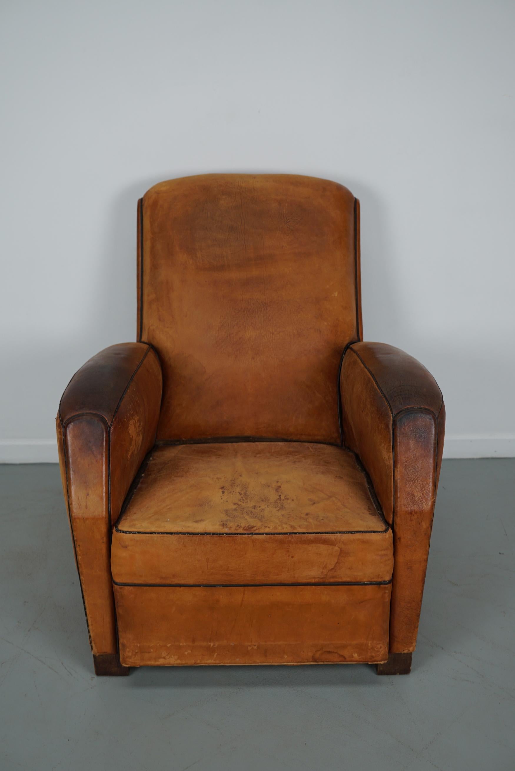 This leather club chair comes from France and was made circa 1950s. It is upholstered with cognac-colored leather and features metal rivets and wooden legs. It remains in a well-used vintage condition with signs of use and age but the chair is still