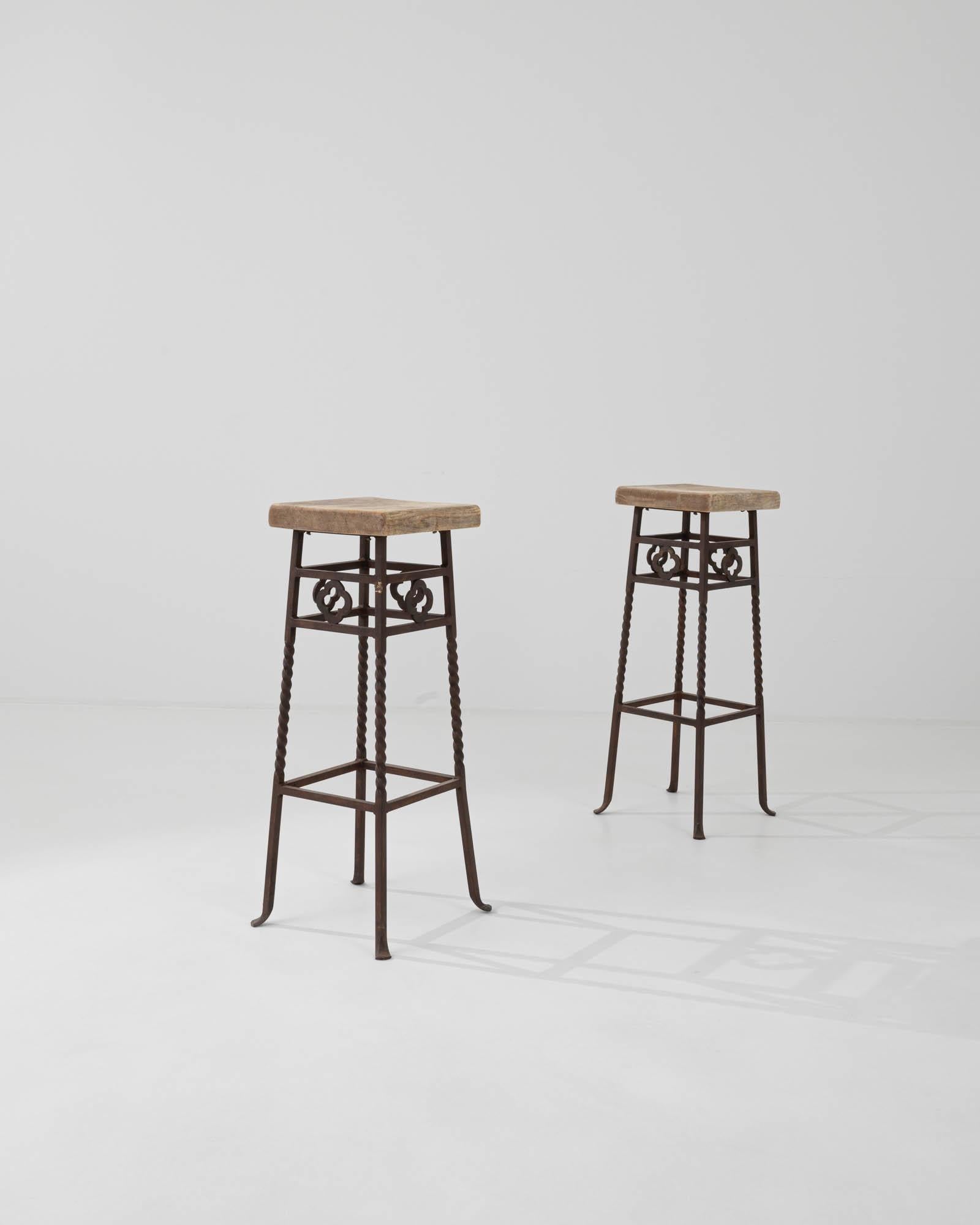 This pair of 20th-century French stools harmoniously blends robust iron with the inviting warmth of wood. Square wooden seats are elevated on a tall iron base that flaunt wrought twists and rosettes, terminating in coquettishly curved feet. The