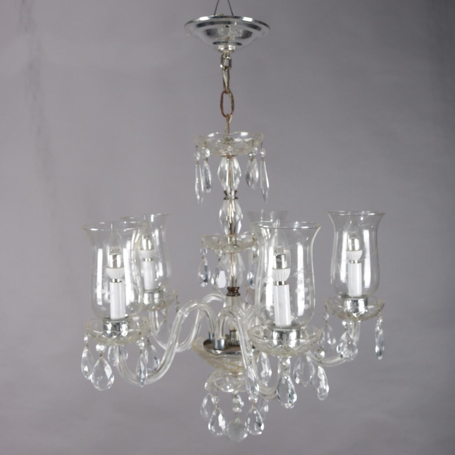 Vintage French chandelier features a chrome frame with central crystal column having s-scroll glass arms terminating in candle lights with etched glass hurricane shades, hanging rock crystal prism highlights throughout, circa 1940.


Measures: 28