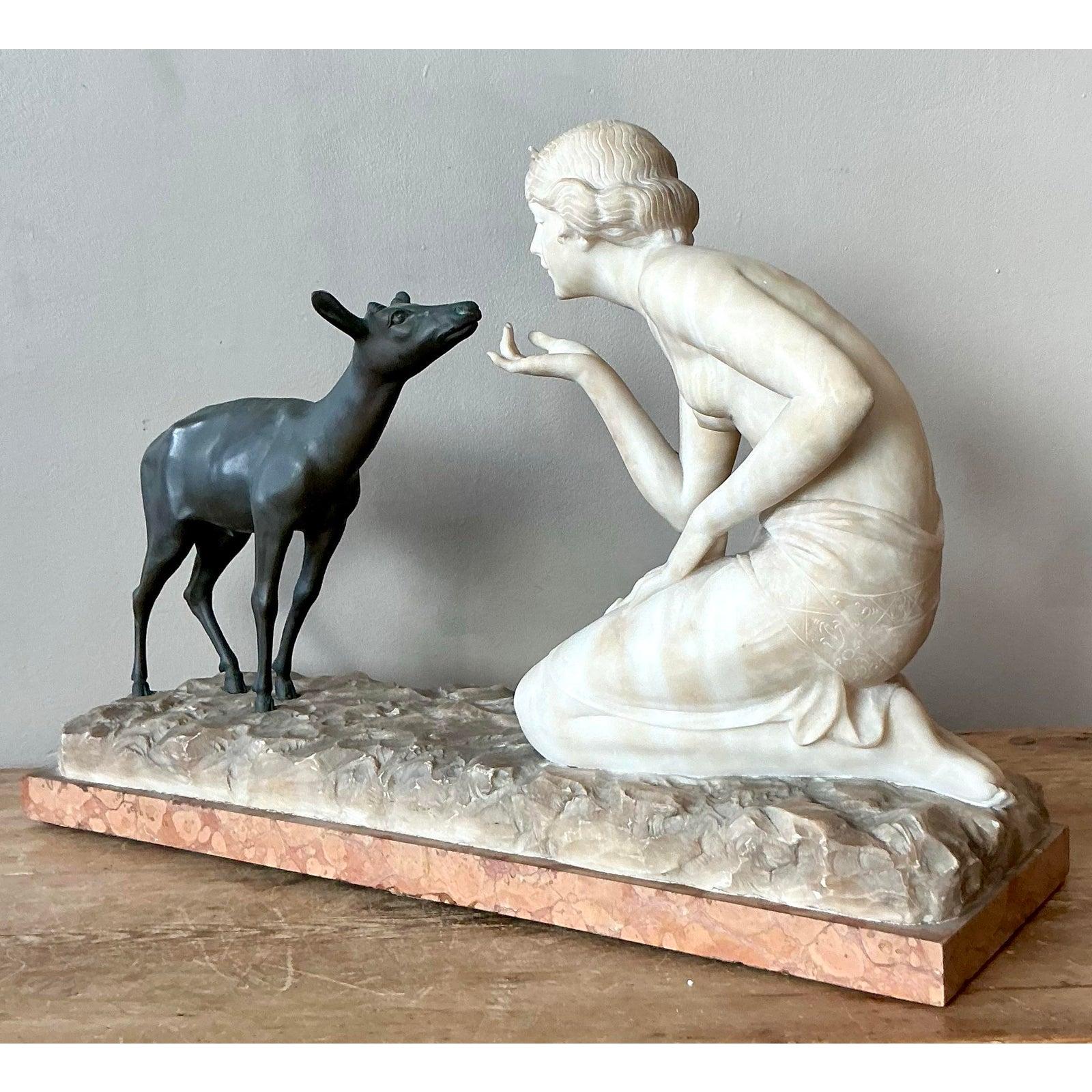 Questa avvincente scultura d'epoca cattura magnificamente un tenero momento tra una figura femminile inginocchiata (che ricorda Diana, la dea romana della caccia) e un piccolo cervo (posteriore). L'opera presenta un sofisticato contrasto di