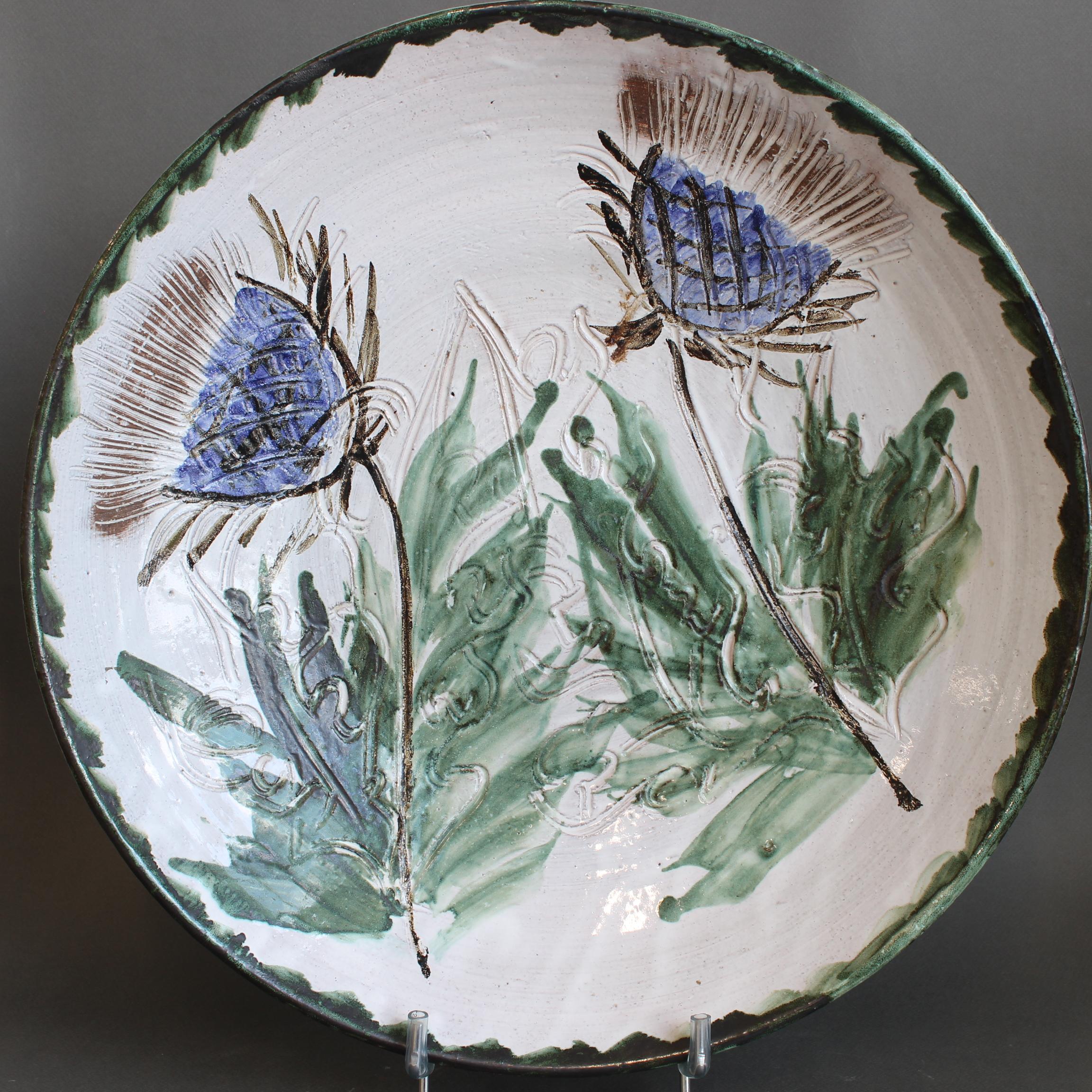 ating from the 1970s. This hand-thrown piece is adorned with delicate blue floral motifs and soft green foliage, all set against a warm, off-white glaze. The loosely painted decoration—signature to Thiry's work—has a lyrical, almost watercolor-like