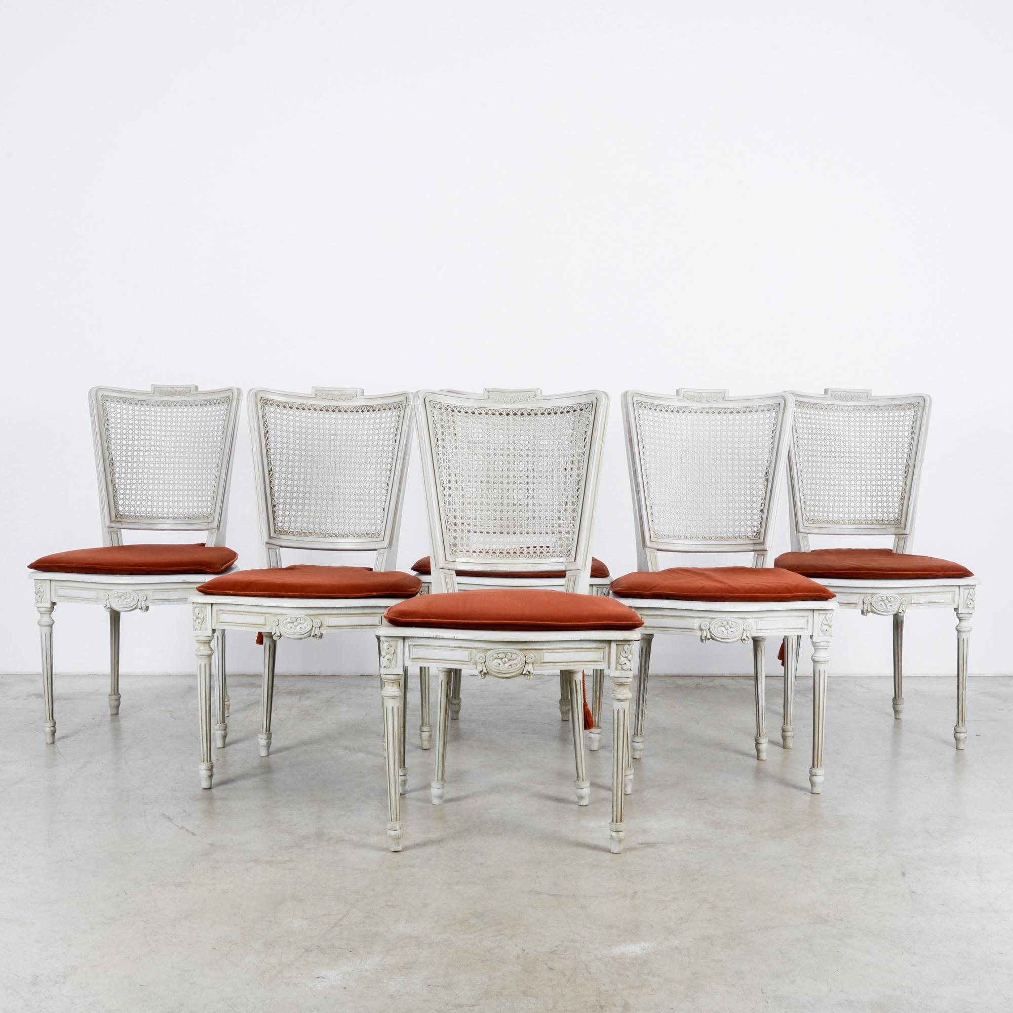 A set of six white wooden chairs with cane backs and red upholstered cushion seats from France, circa 1960. Louis XVI inspired design — with floral motif appearing in the crest, crowning the legs, and as centerpiece of the apron — is an understated