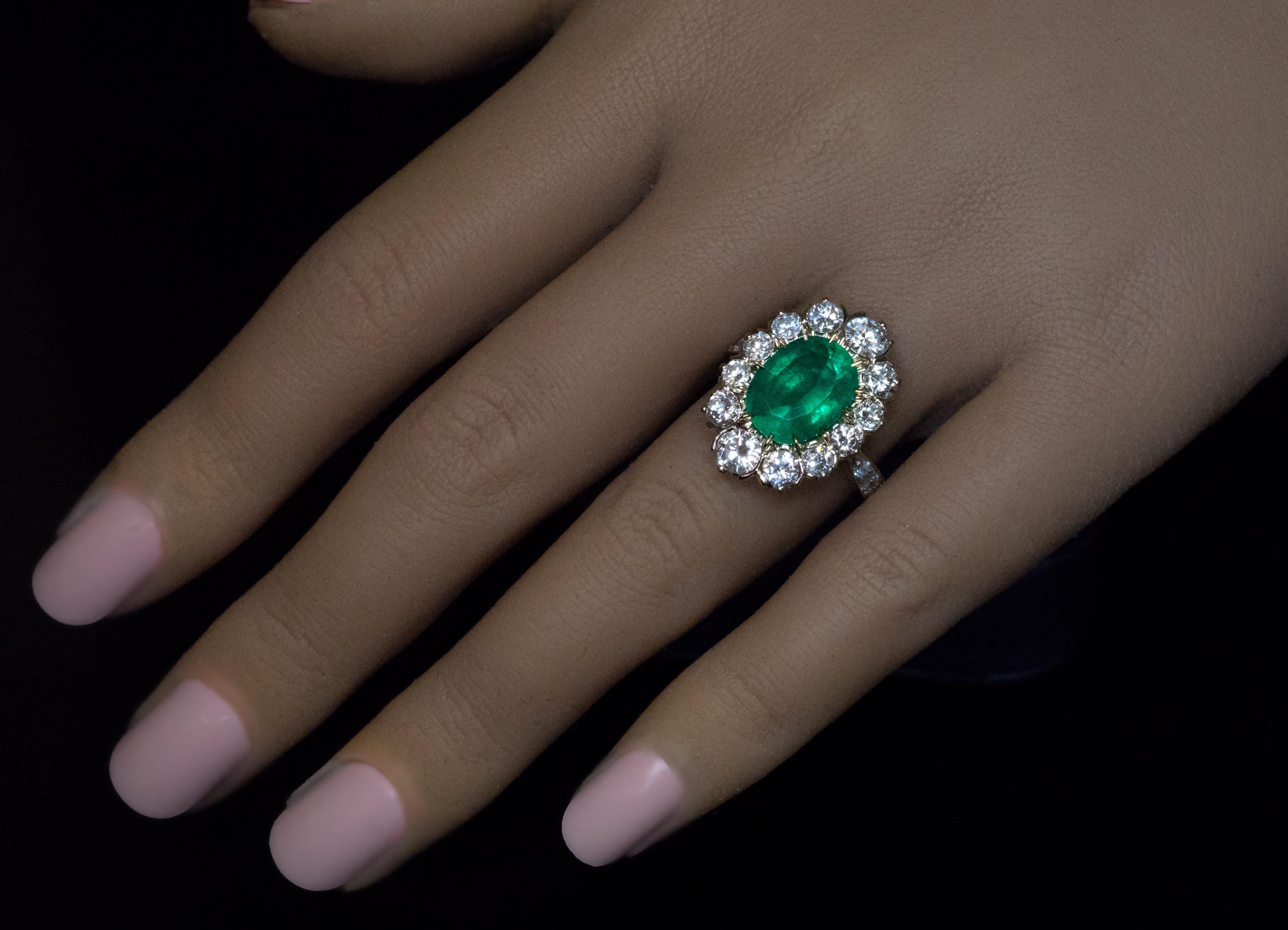 France, c. 1950s  This impressive cluster ring is finely crafted in 18K yellow and white gold. The ring is centered with an oval 3.64 ct emerald of a vivid bluish green color. The emerald is surrounded by bright white brilliant cut diamonds (F-G