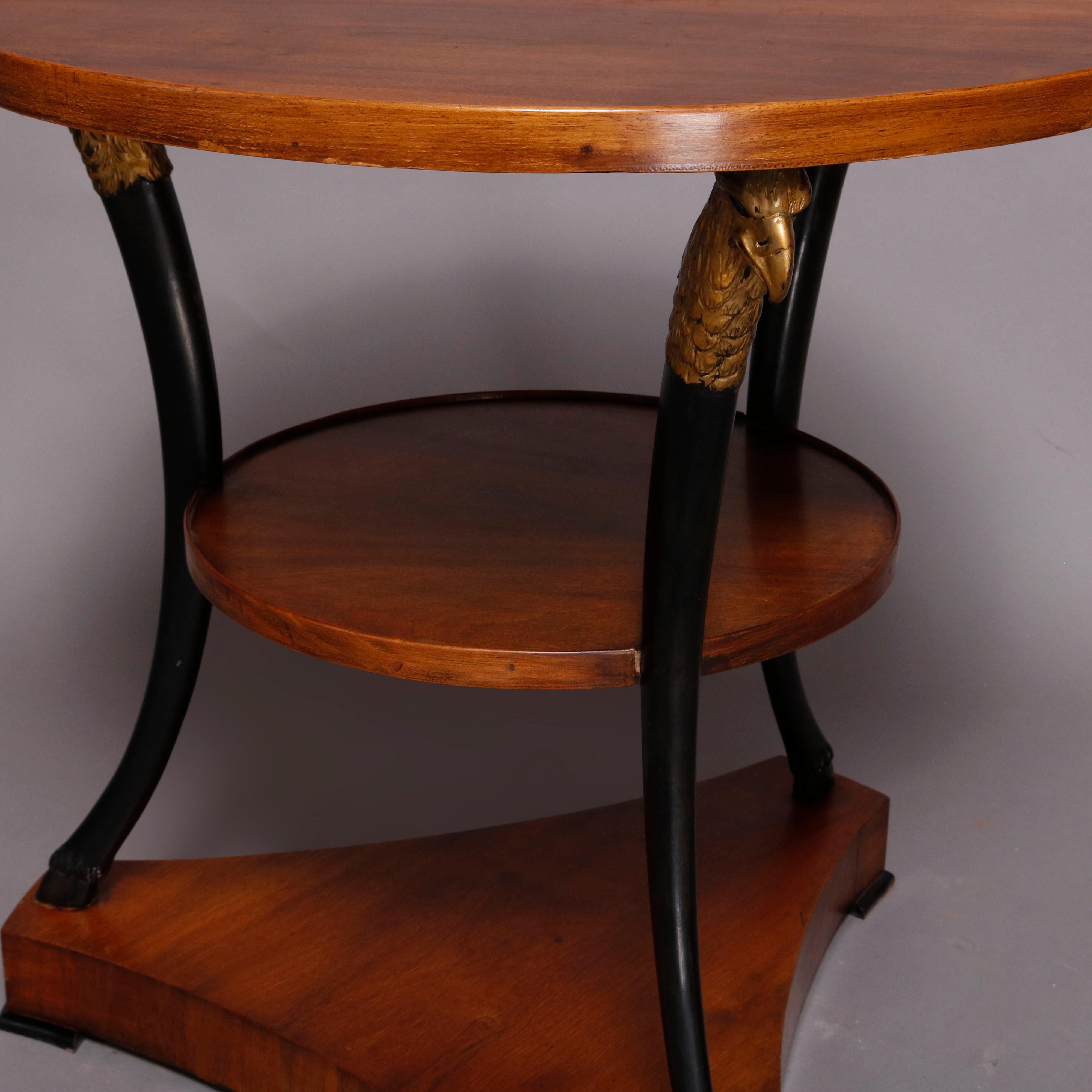 A vintage French Empire figural lamp stand offers mahogany construction with round top surmounting lower round display supported by three convex ebonized supports each having a gilt falcon head and terminating in a hoof foot seated on triangular